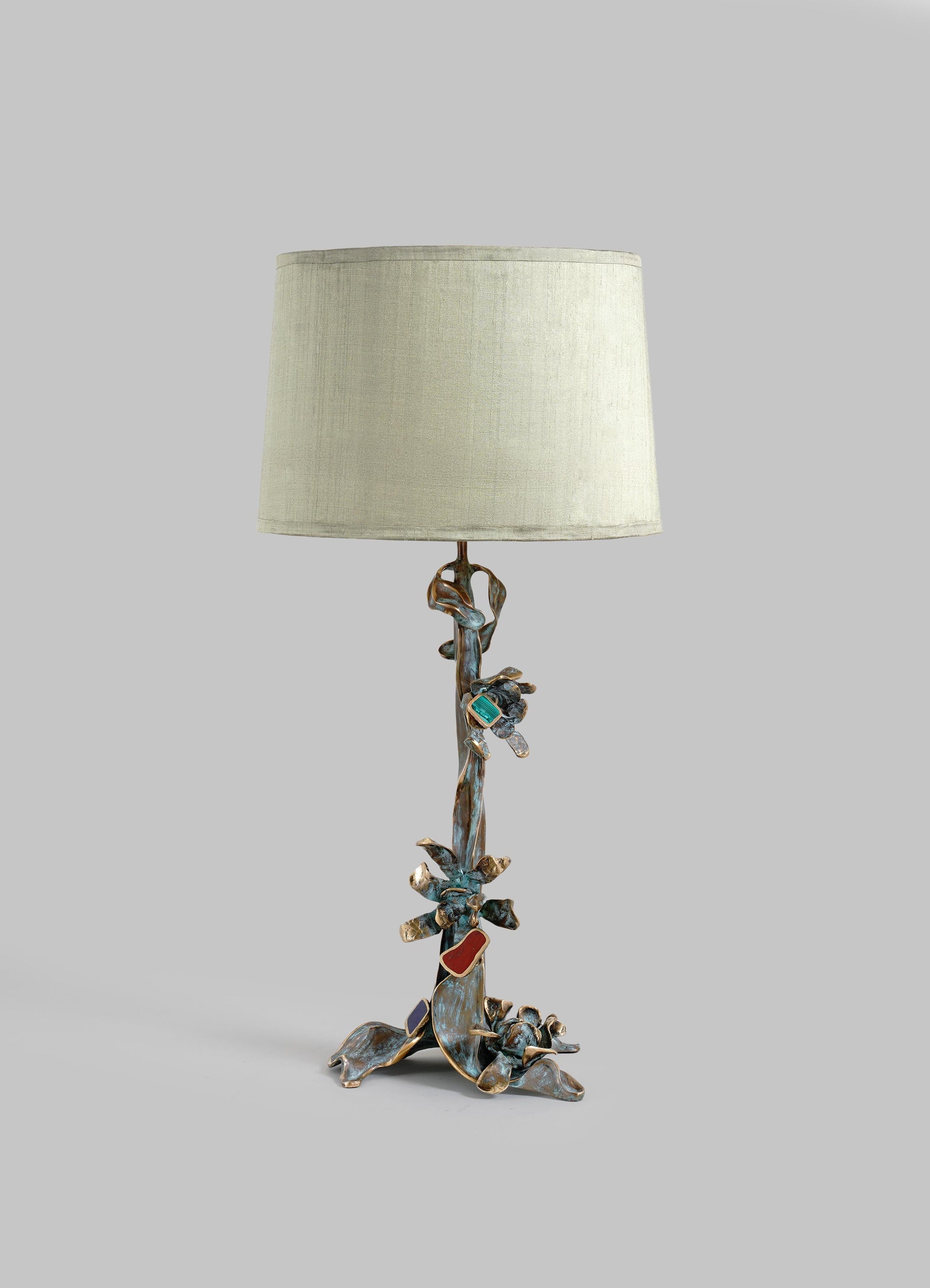 Decorative table lamp with a sculptural metal base resembling a natural branch or vine, finished in bronze and featuring colorful glass or semi-precious stone accents, paired with a neutral-colored fabric lampshade.