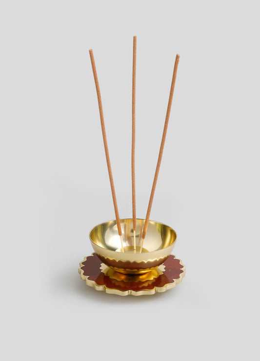 A decorative incense holder with a polished gold interior bowl, featuring a scalloped edge in red and gold tones, and three long wooden incense sticks inserted vertically.