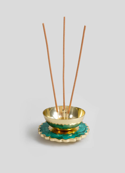 A decorative incense holder with a gold-colored metal bowl resting on a scalloped ceramic base in teal and gold tones. Four wooden incense sticks are inserted into the bowl.