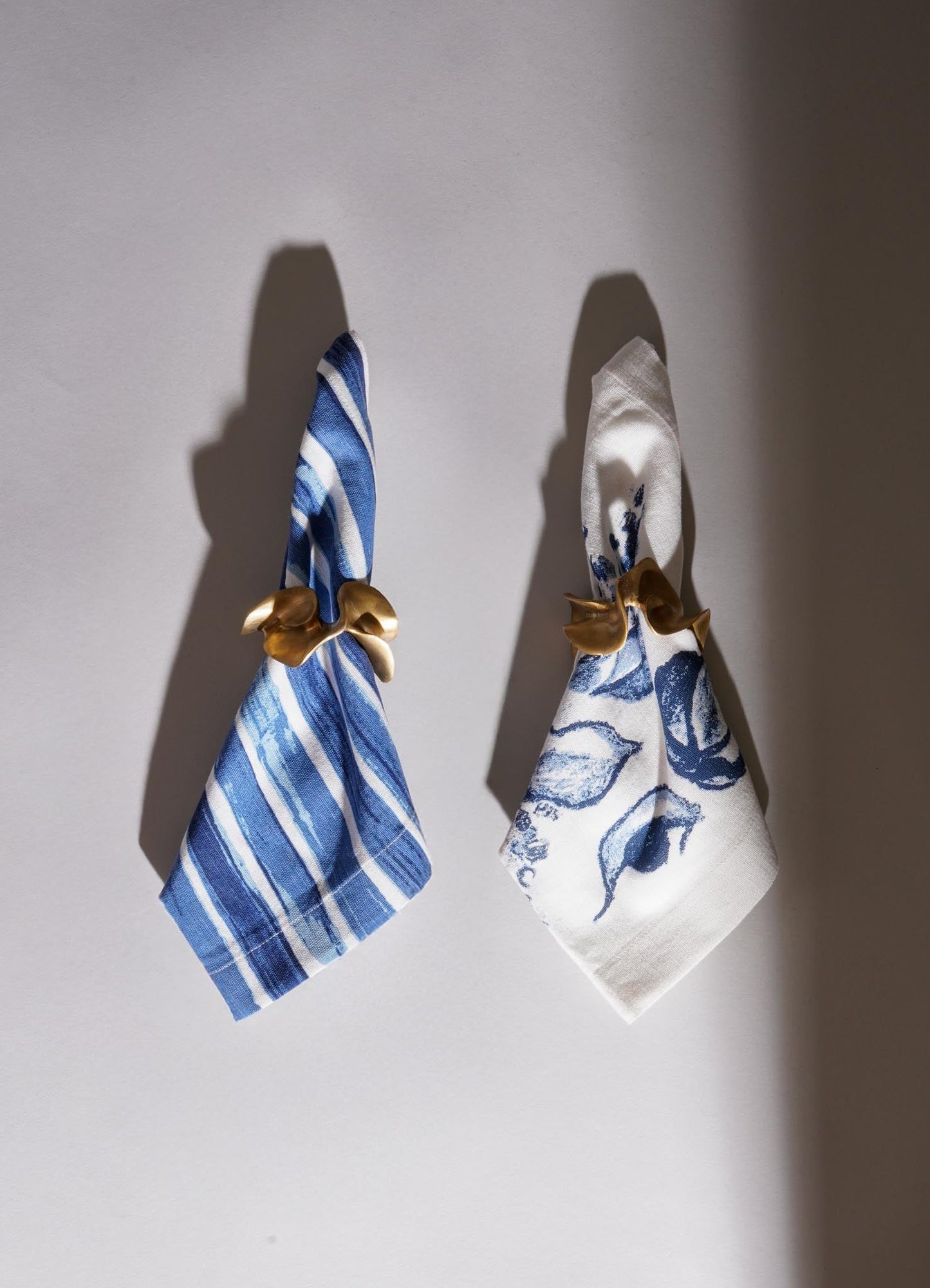 Set of two fabric napkins, one with blue and white striped pattern and the other with a blue floral print on white, held by gold-toned napkin rings shaped like abstract leaves. The napkins are hanging on a plain wall with shadows cast by the rings and fabric.