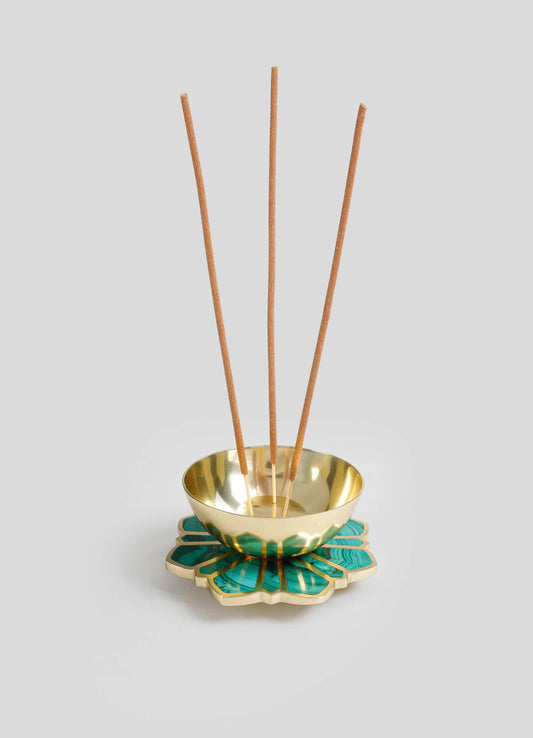 A decorative incense holder featuring a gold metal bowl with a polished finish, resting on a ceramic base with a teal and gold geometric floral motif. Four wooden incense sticks are placed inside the bowl.