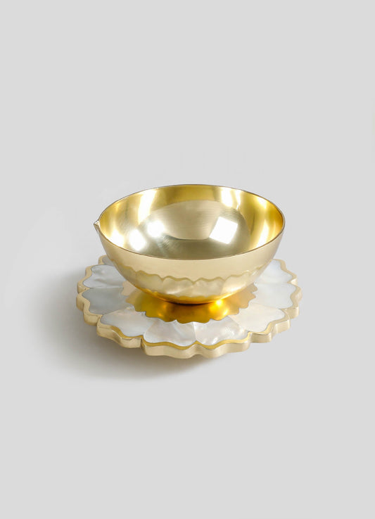 A gold-colored metal bowl with a smooth reflective surface sits atop a white ceramic or porcelain saucer with scalloped edges and gold trim. The design is simple and elegant, with a focus on metallic and ceramic materials.