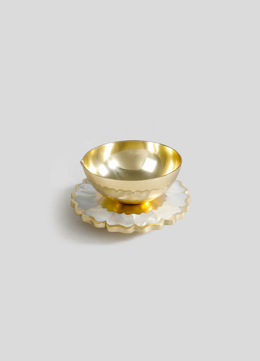 A gold-toned metal bowl with a polished, reflective surface sits atop a layered shell-shaped tray with white and cream hues and wavy edges.