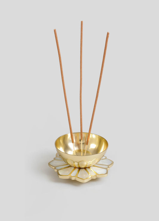 A decorative brass bowl with a polished gold finish placed on a white floral-shaped ceramic tray, holding three tall wooden incense sticks. The design combines luxury metallic elements with handcrafted details.