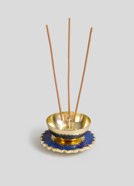 A gold-toned metal bowl with a reflective interior, placed on a decorative blue and gold ceramic dish with a scalloped edge, accompanied by three wooden reed sticks.