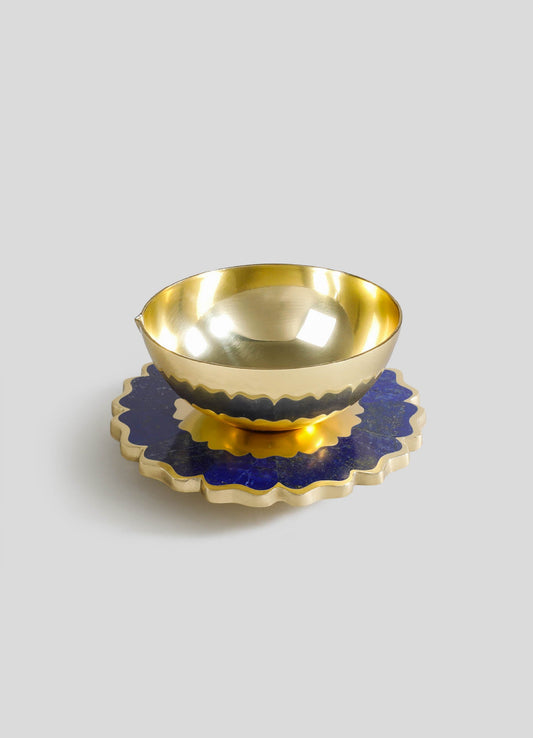 Polished gold-plated metal bowl with a smooth, reflective surface, placed on a dark blue and gold scalloped ceramic dish with a textured pattern.