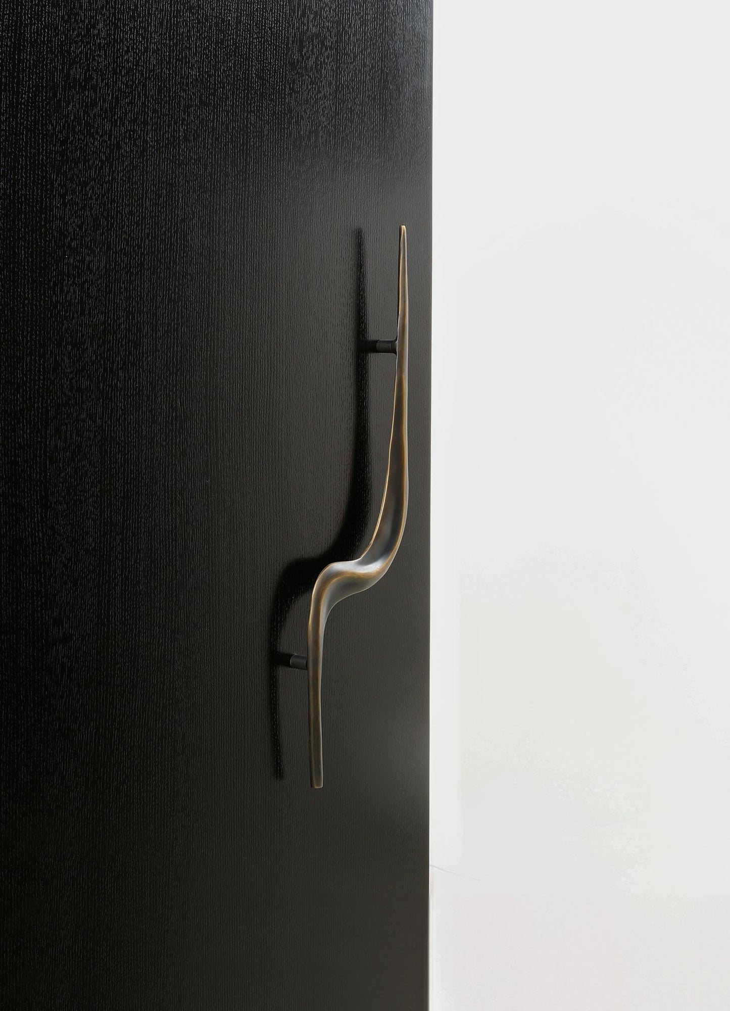A modern wall-mounted coat hook with a sleek, curved brass finish, attached to a black textured wall panel, contrasting against a plain white background.