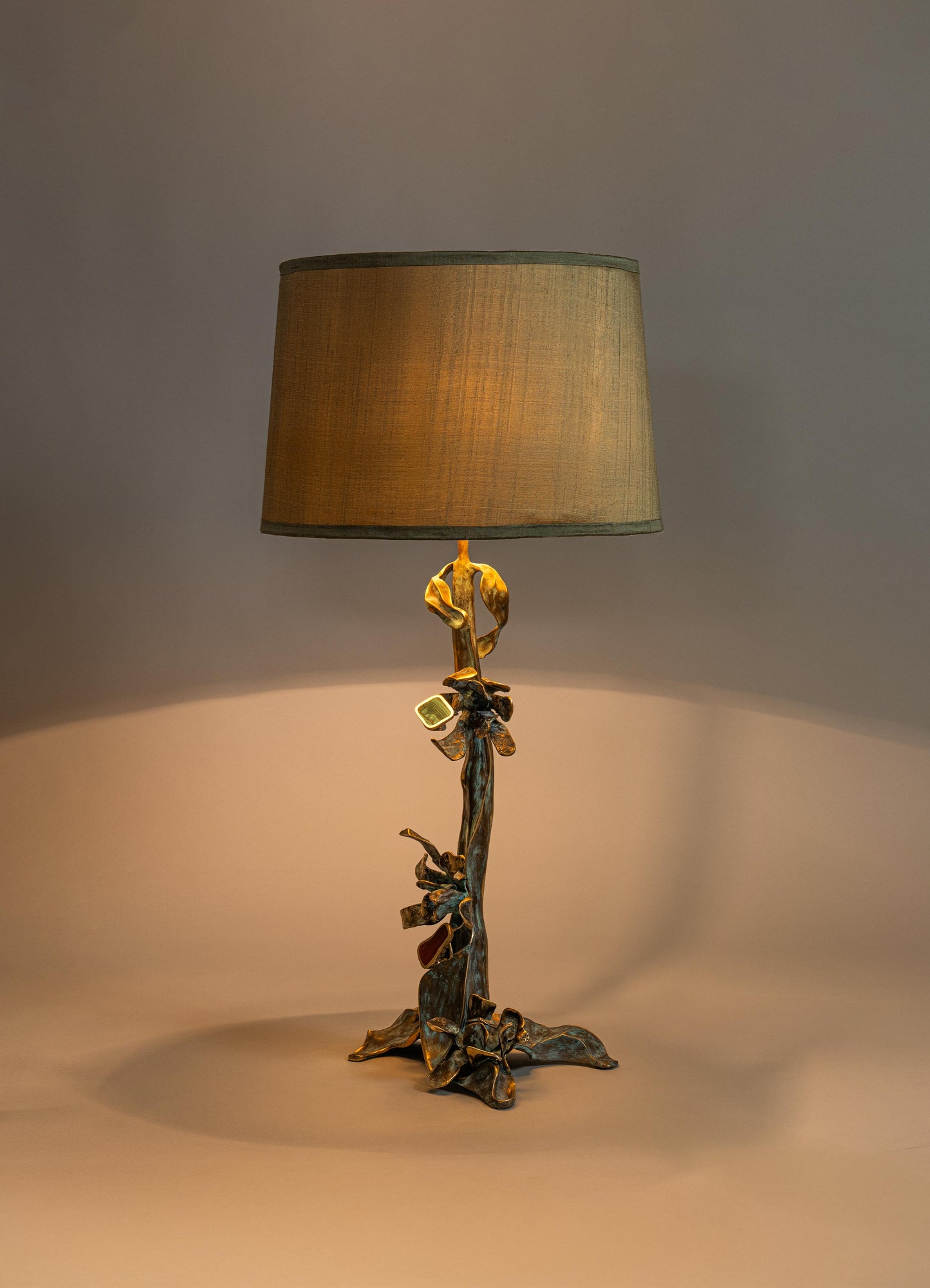 A table lamp with a sculptural, nature-inspired metal base featuring a bronze finish and intricate leaf details, topped with a large cylindrical fabric lampshade in beige.