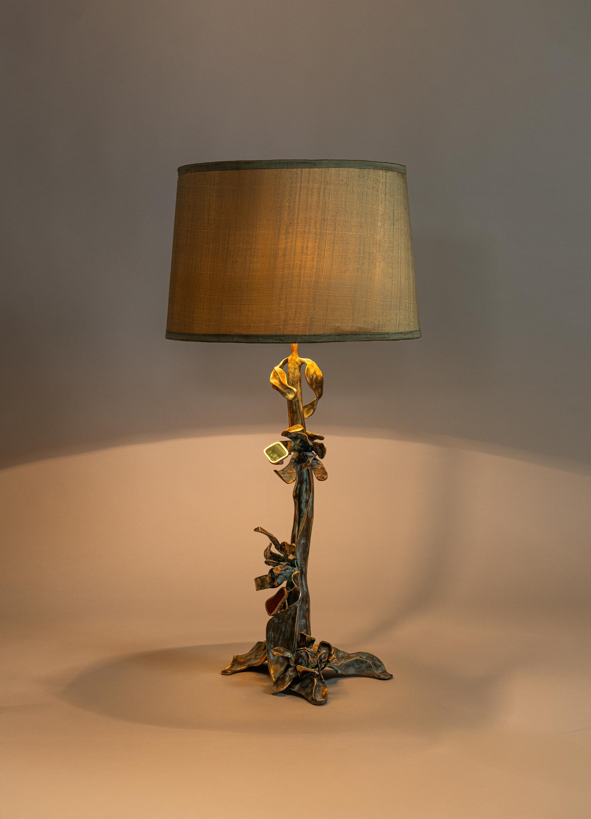 A table lamp with a sculptural, nature-inspired metal base featuring a bronze finish and intricate leaf details, topped with a large cylindrical fabric lampshade in beige.