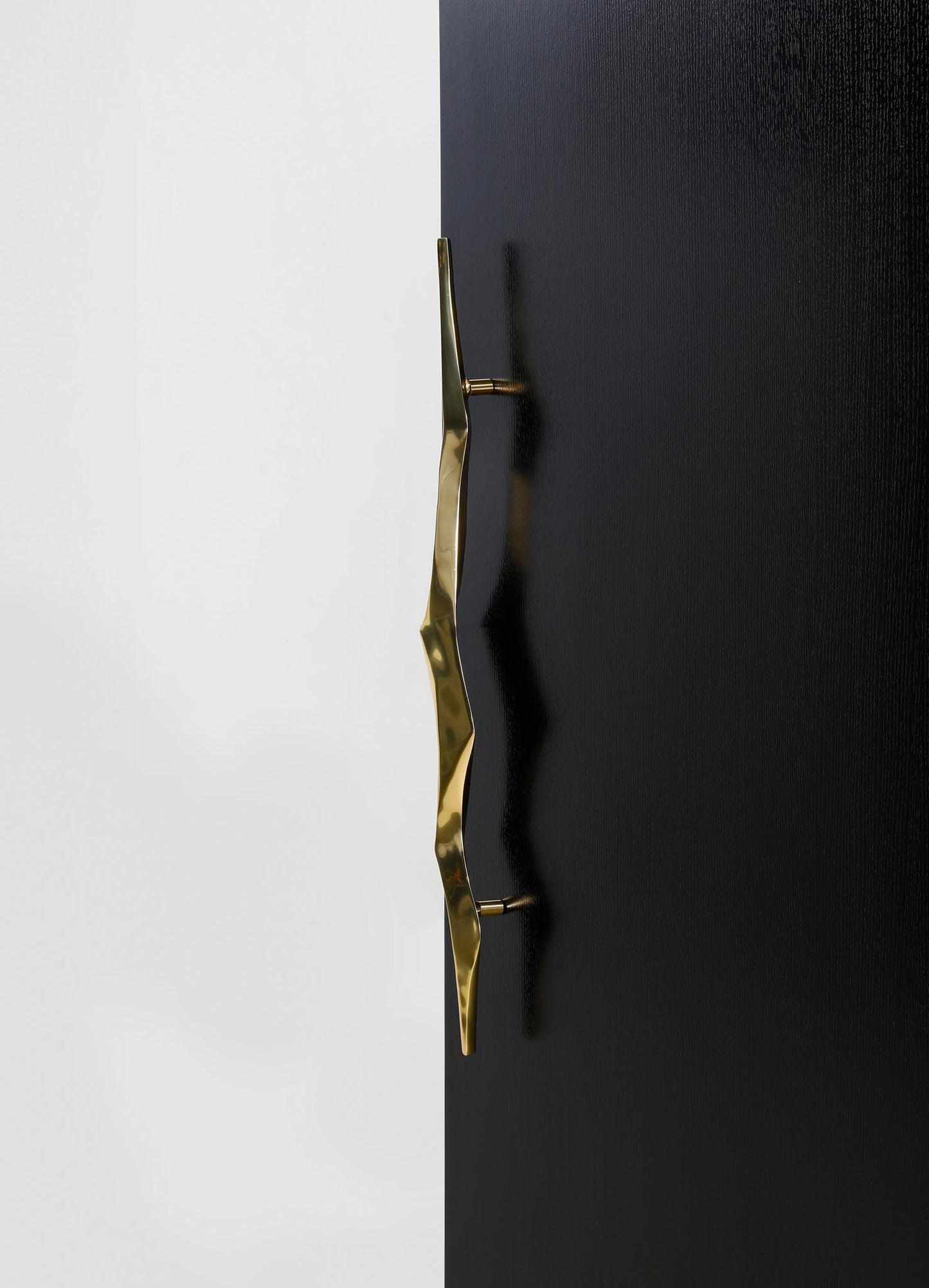A modern brass door handle with a wavy, textured design mounted on a black door, contrasting with a white wall background.