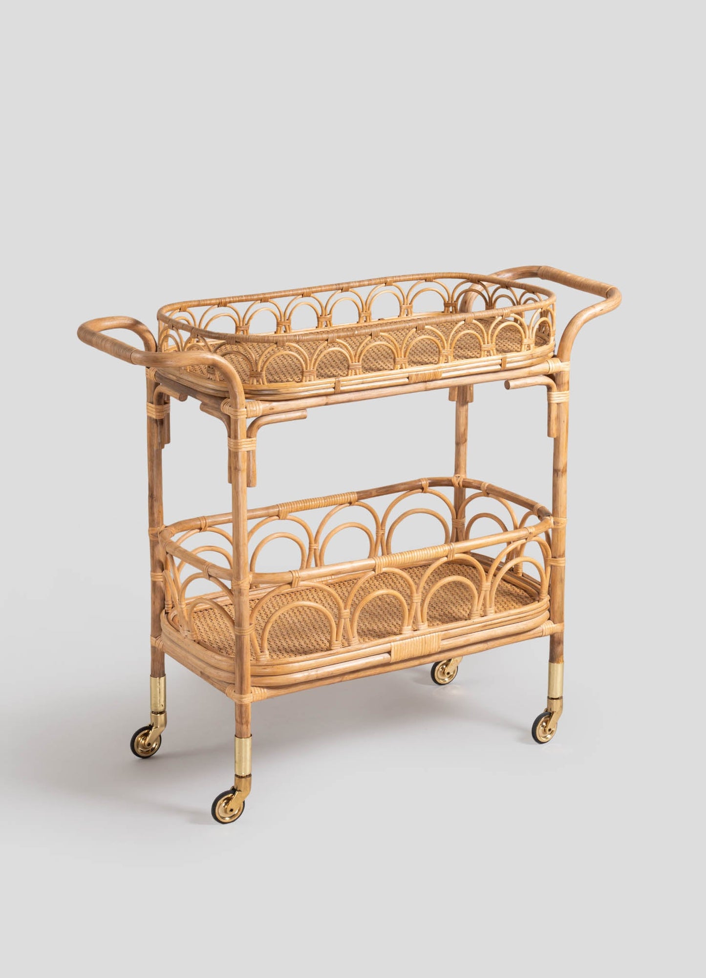 Wicker bar cart with two tiers featuring rounded arch motifs and cane-weaved shelf surfaces, mounted on brass-capped caster wheels. The cart has curved rattan handles and a natural light wood finish, designed in a rustic or vintage style.