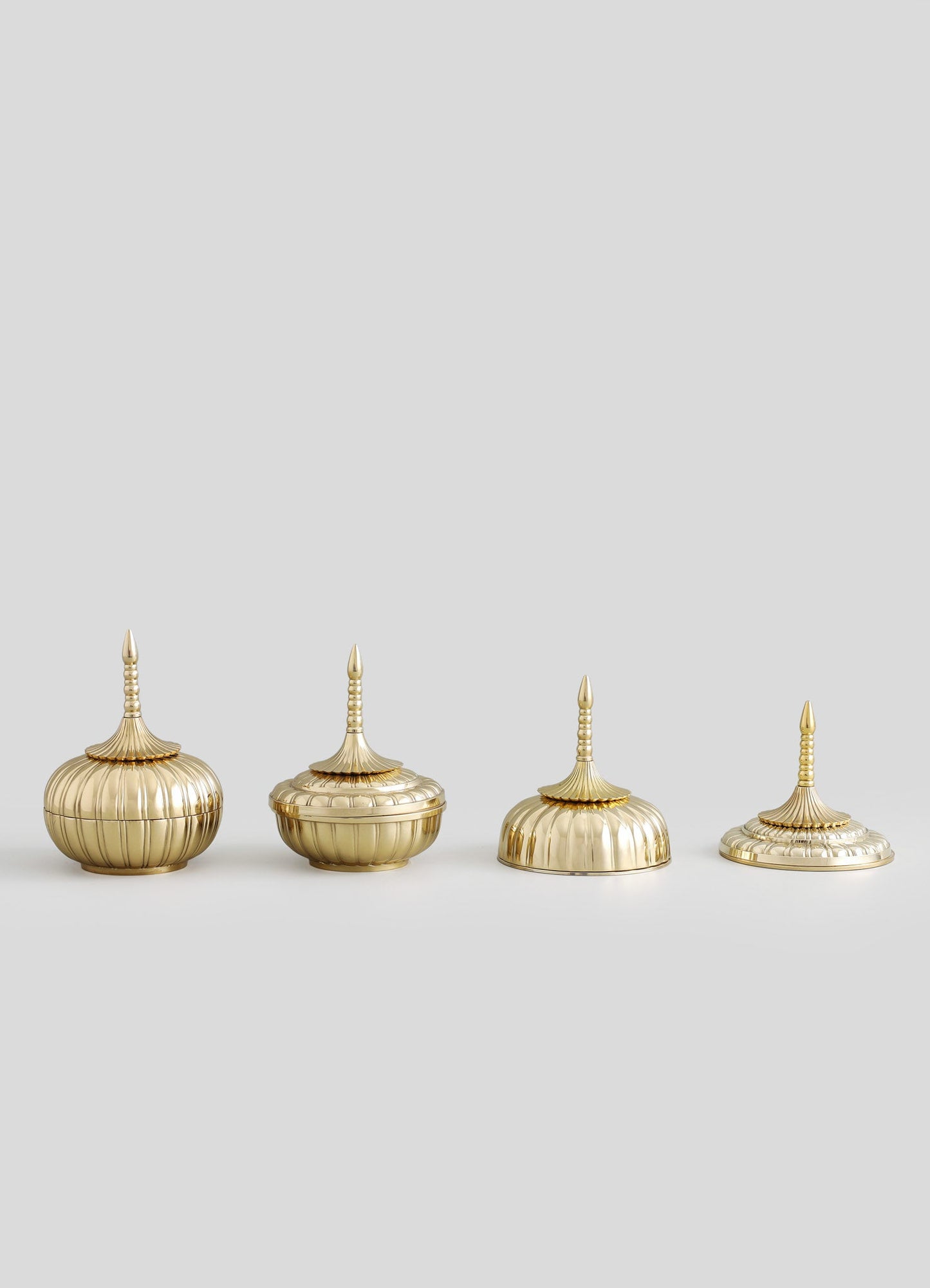 Four decorative brass containers with ribbed, rounded bodies and tall, pointed lids featuring layered, spindle-like finials, arranged in a line against a plain background.