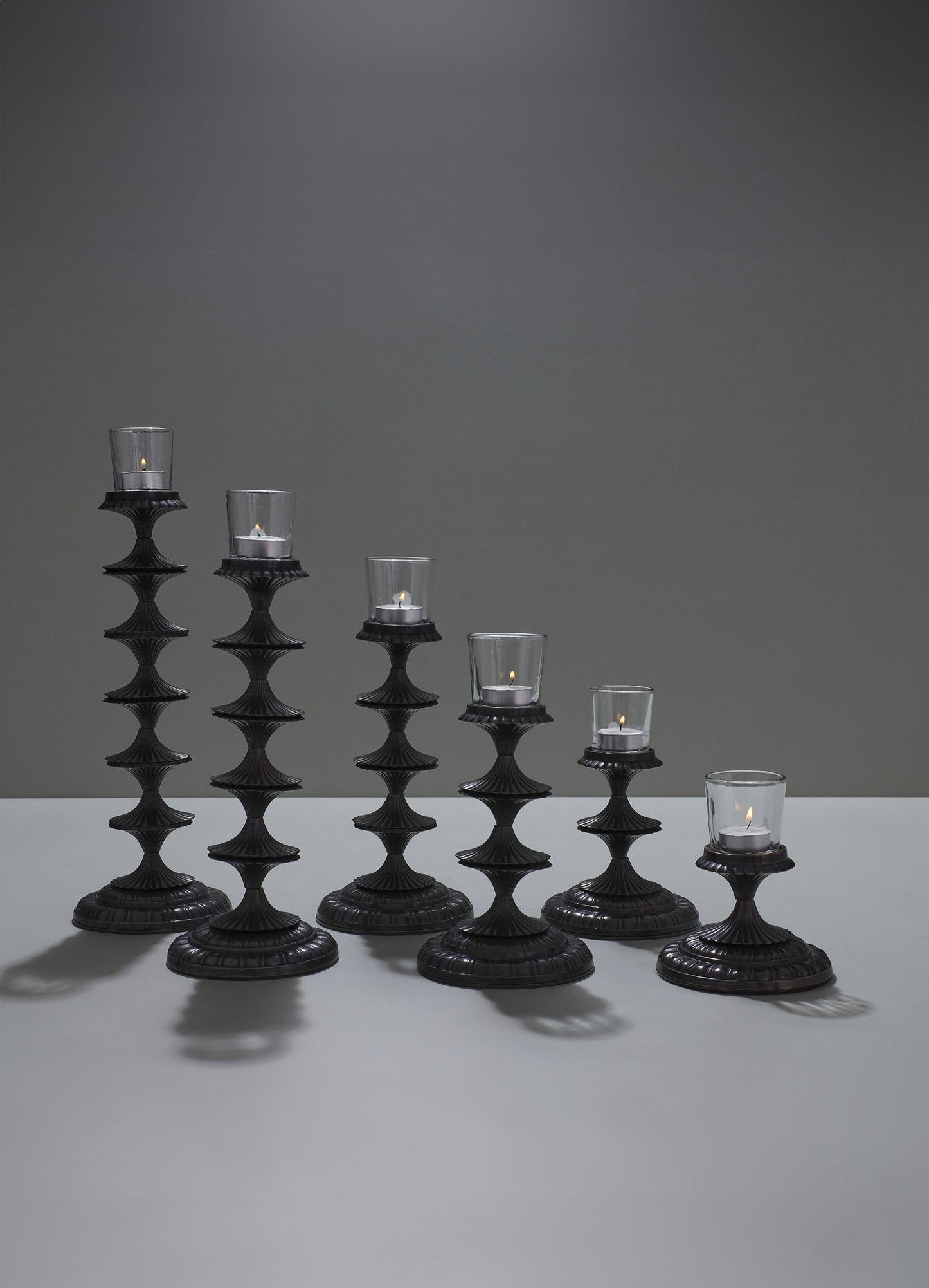 Set of six black candlesticks with carved, tiered bases and transparent glass hurricane protectors, each holding a white tea light candle, arranged on a light gray surface against a darker gray background.