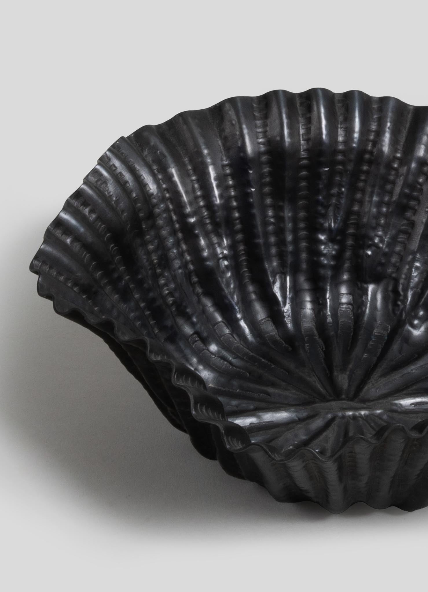 Black shell-shaped bowl made of glossy material with a ribbed, textured surface and a scalloped edge, set against a plain light background.