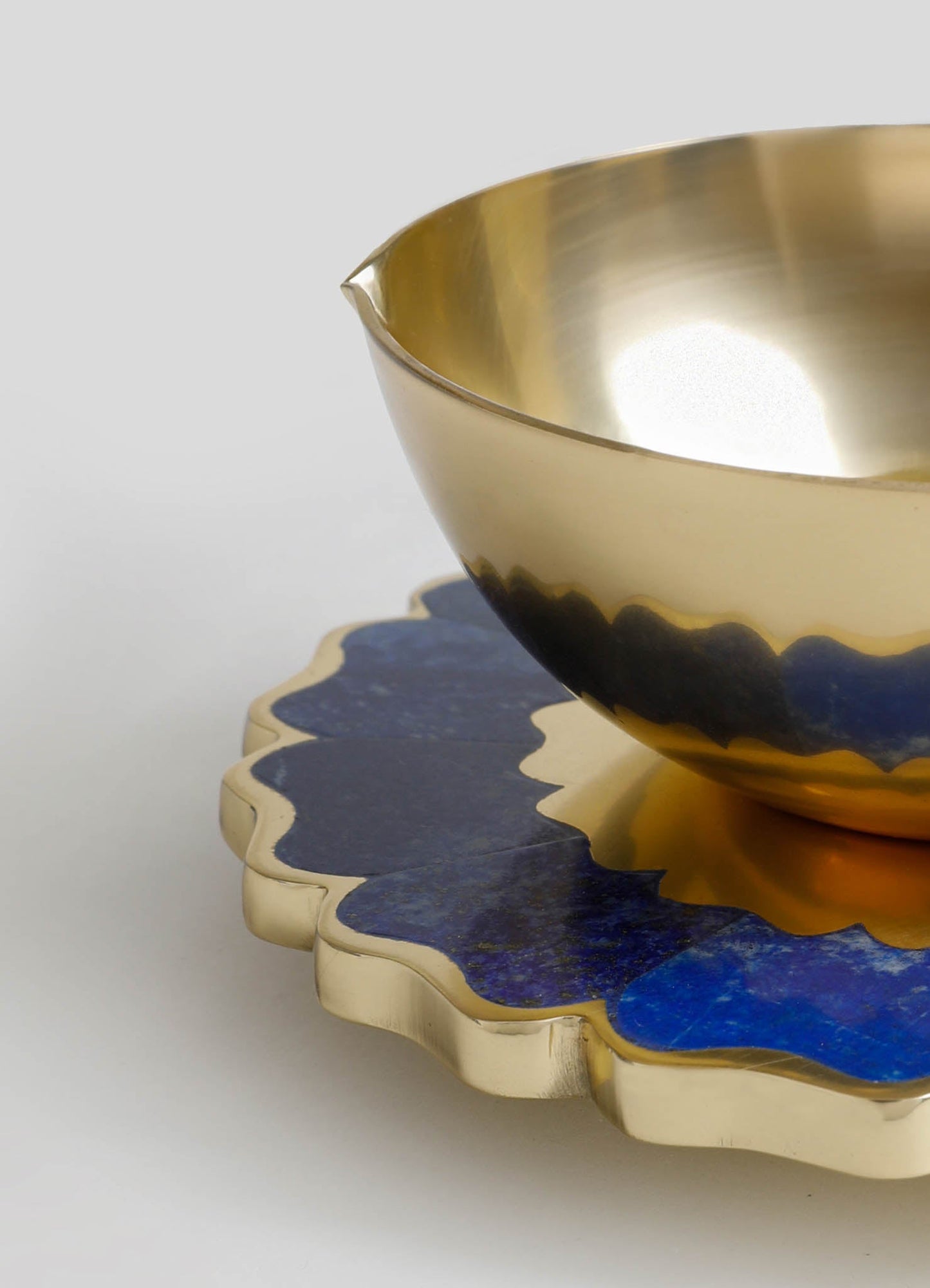 A decorative brass bowl with a polished gold finish sits on a scalloped ceramic dish featuring a blue and gold abstract pattern. The combination of materials and colors creates a luxurious and contemporary aesthetic.