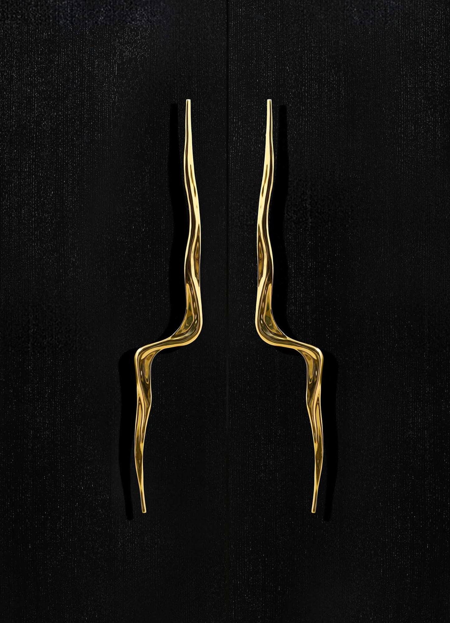 Gold-toned sculptural door handles with wavy, elongated shapes mounted on a black door surface.