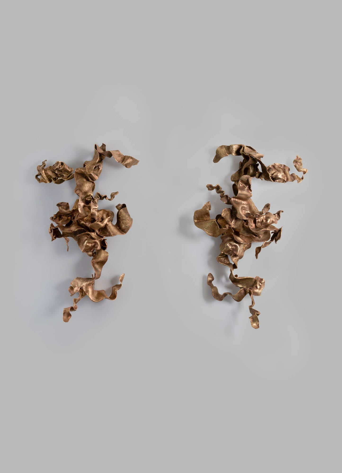 Two abstract wall sculptures made of metallic copper-colored material with organic, ruffled, and twisted forms are displayed against a light gray background. The sculptures have a textured surface and resemble natural, flowing shapes.