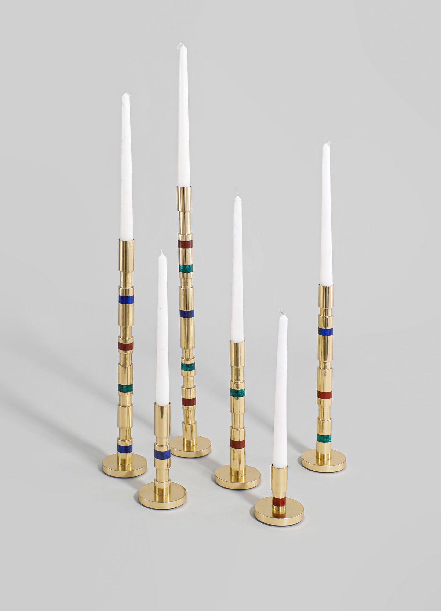 Set of eight tall candles in white wax with gold metal holders featuring colorful geometric accents in blue, red, and green, arranged on a light neutral background.