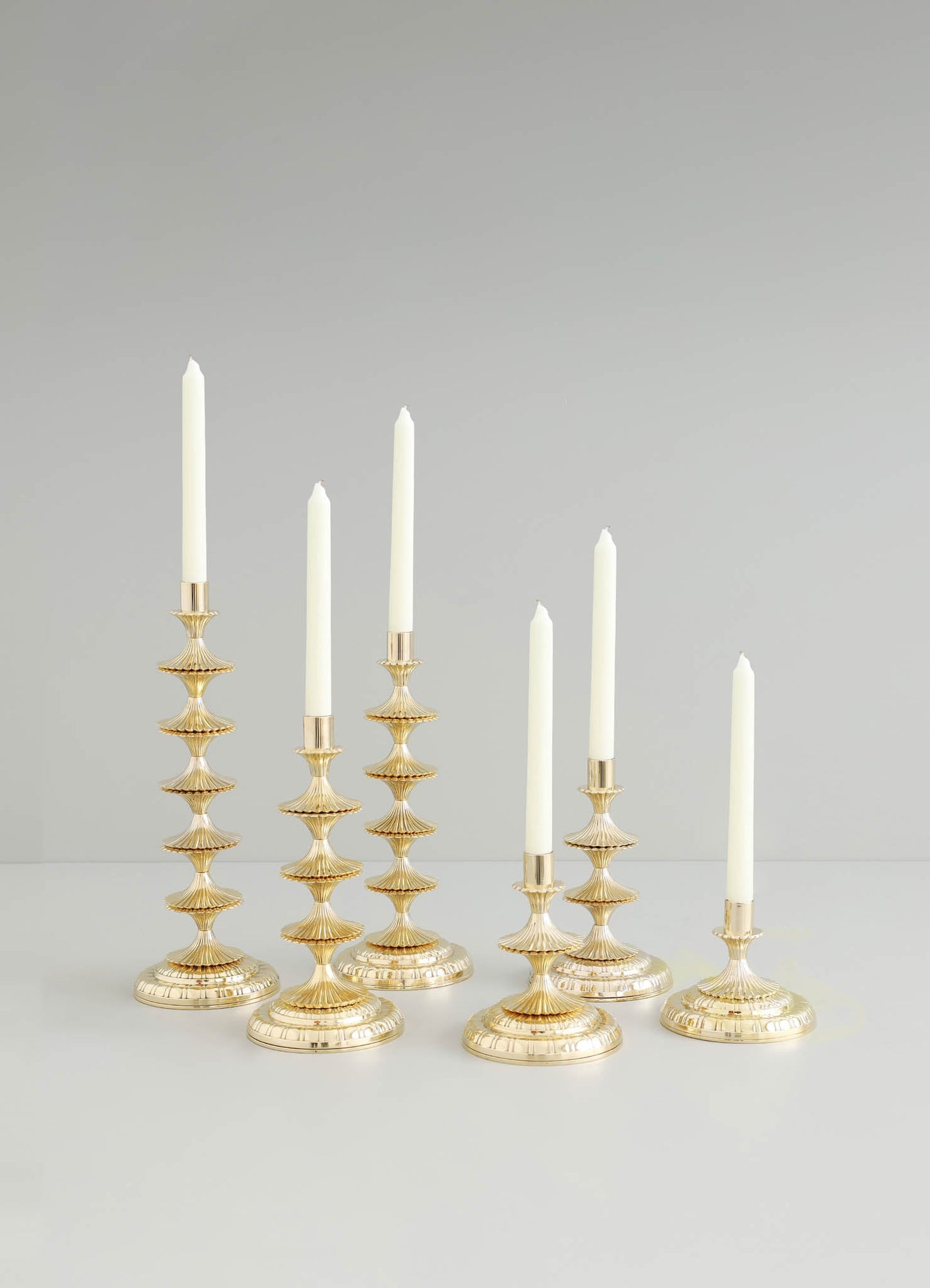 Set of six brass candle holders with tiered, fluted design and a polished gold finish, each holding a white taper candle. The candle holders feature a classical style with rounded bases, arranged on a white surface against a plain background.