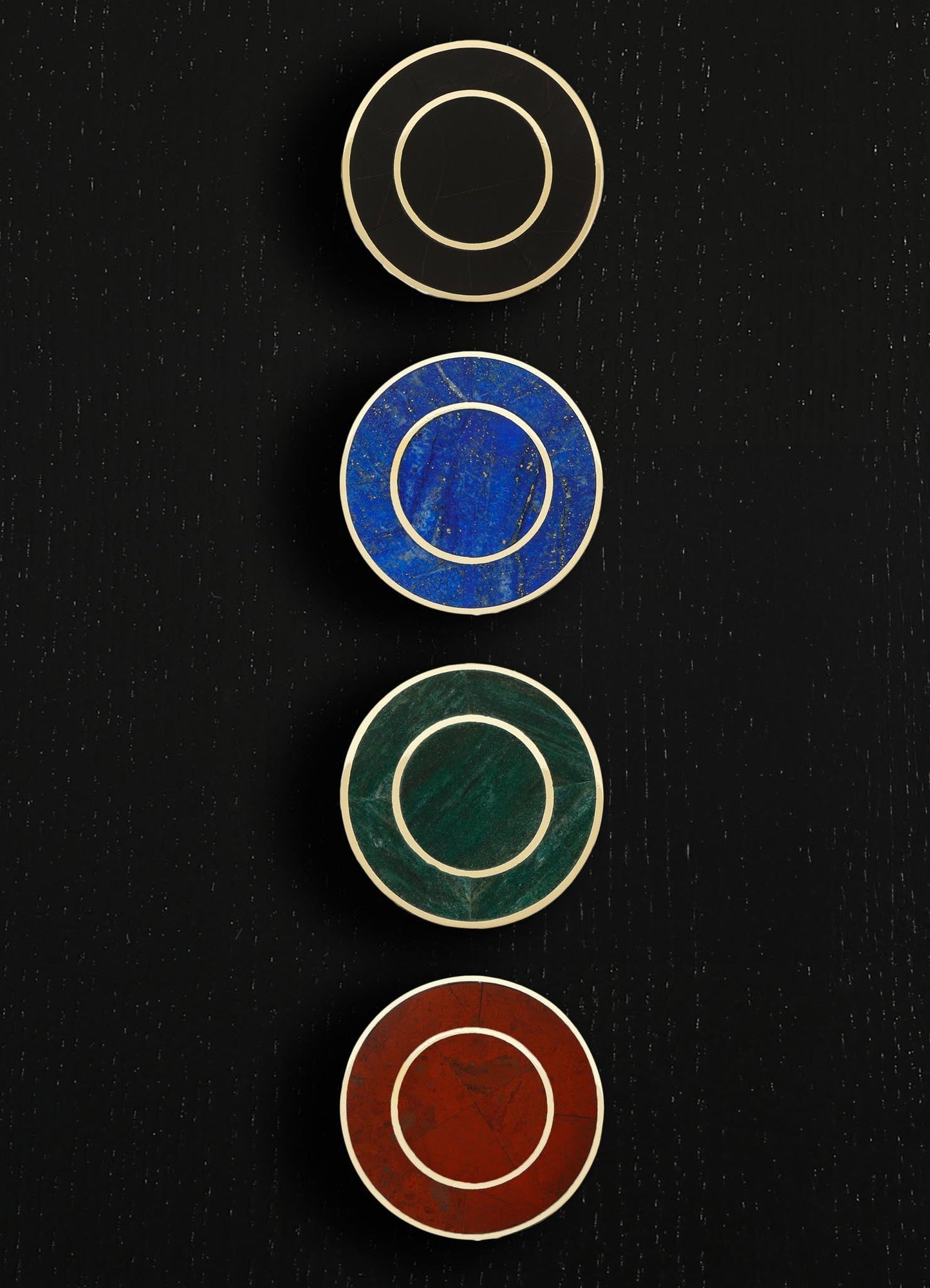 Four circular decorative wall pieces featuring different materials and colors on a black textured background. The top circle appears to be black with a light-colored ring, followed by a blue circle with a speckled pattern, a green marble-like circle, and a red-brown marble-like circle.