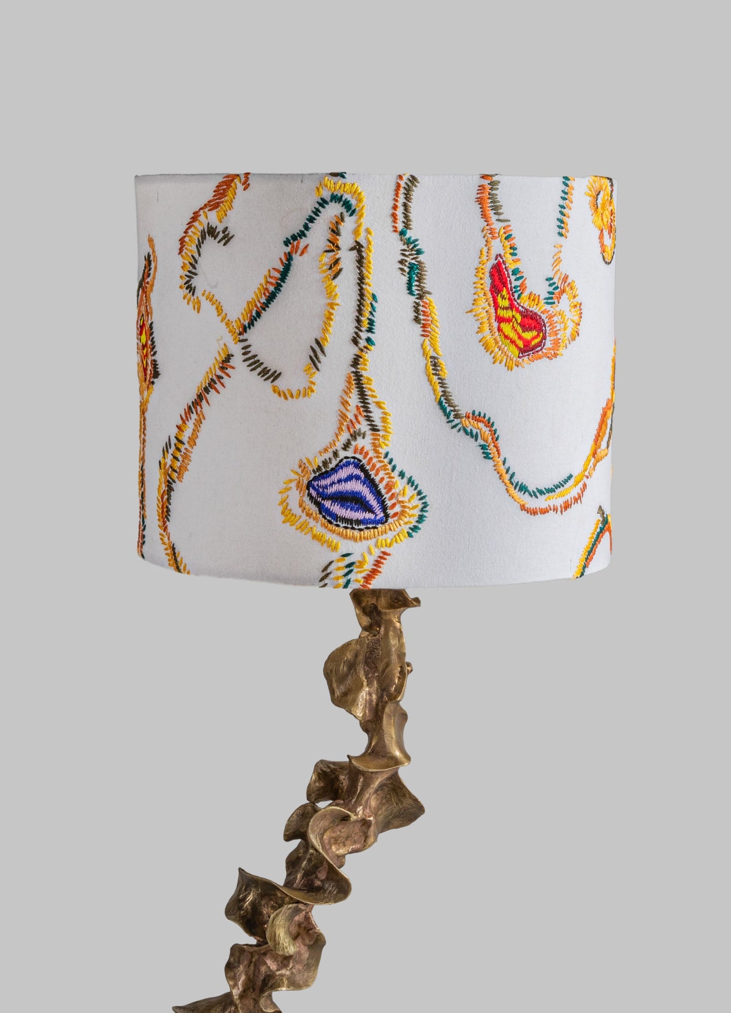 Table lamp with embroidered fabric shade featuring colorful floral motifs on a white background, and a sculptural bronze base with organic, irregular shapes.