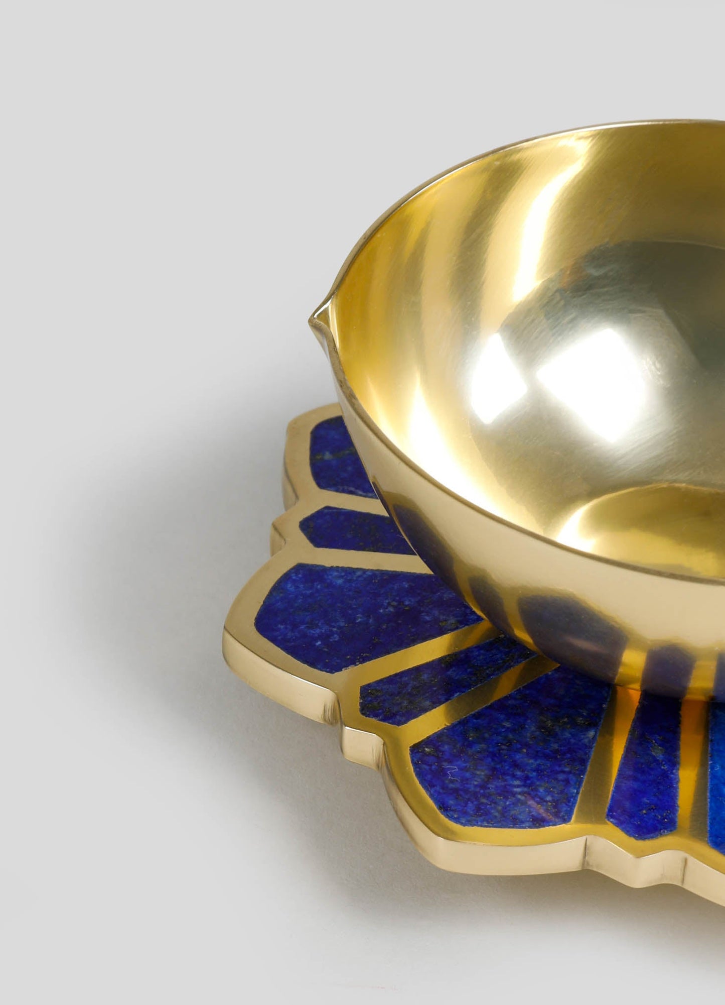 A gold-colored metal bowl with a reflective finish and a smooth, rounded shape. It rests on a decorative base featuring blue and gold geometric patterns with a scalloped edge.