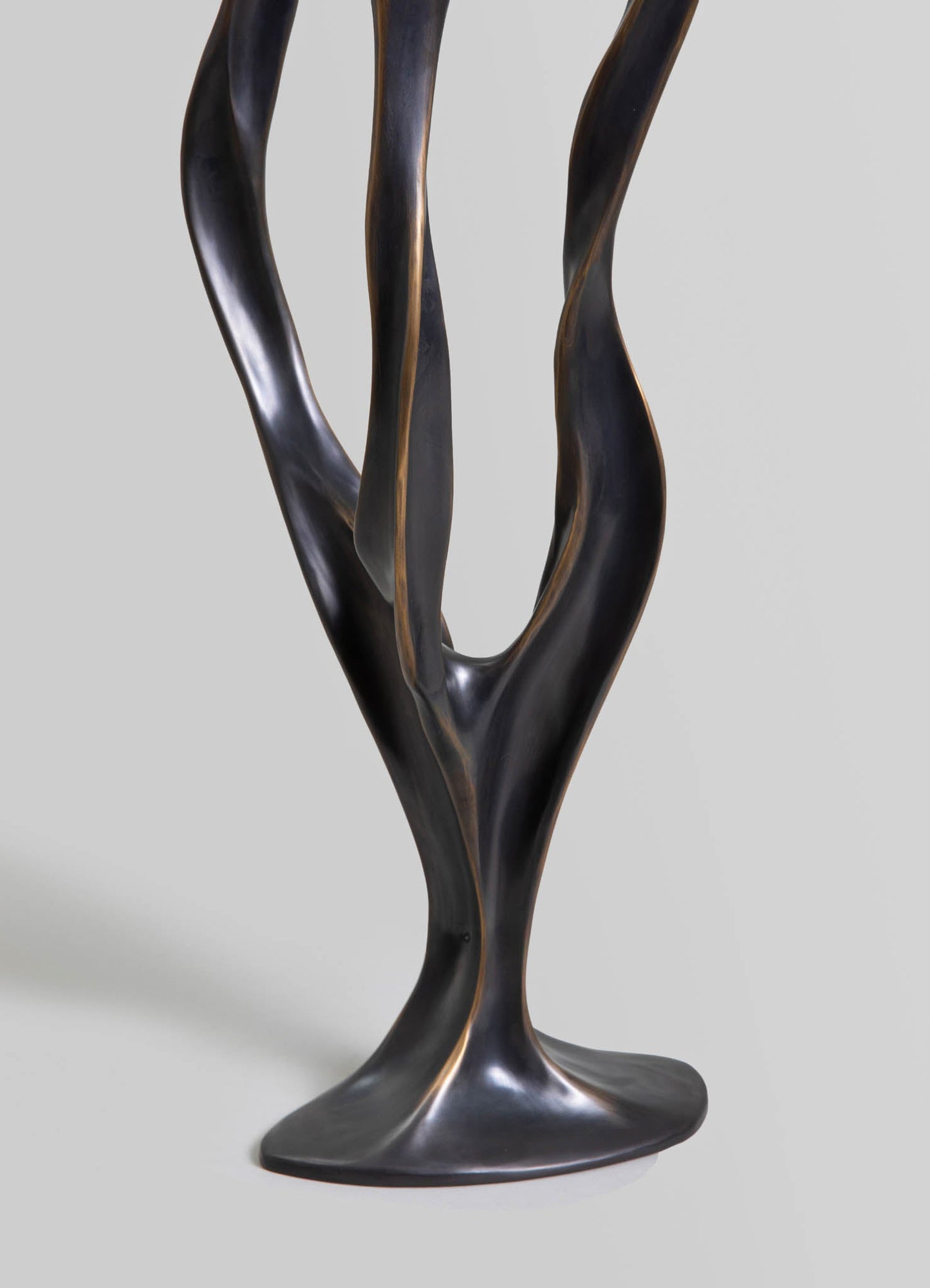 Sculptural metal table base with twisted, organic-shaped arms in a dark, matte finish with subtle bronze accents, set against a plain background.