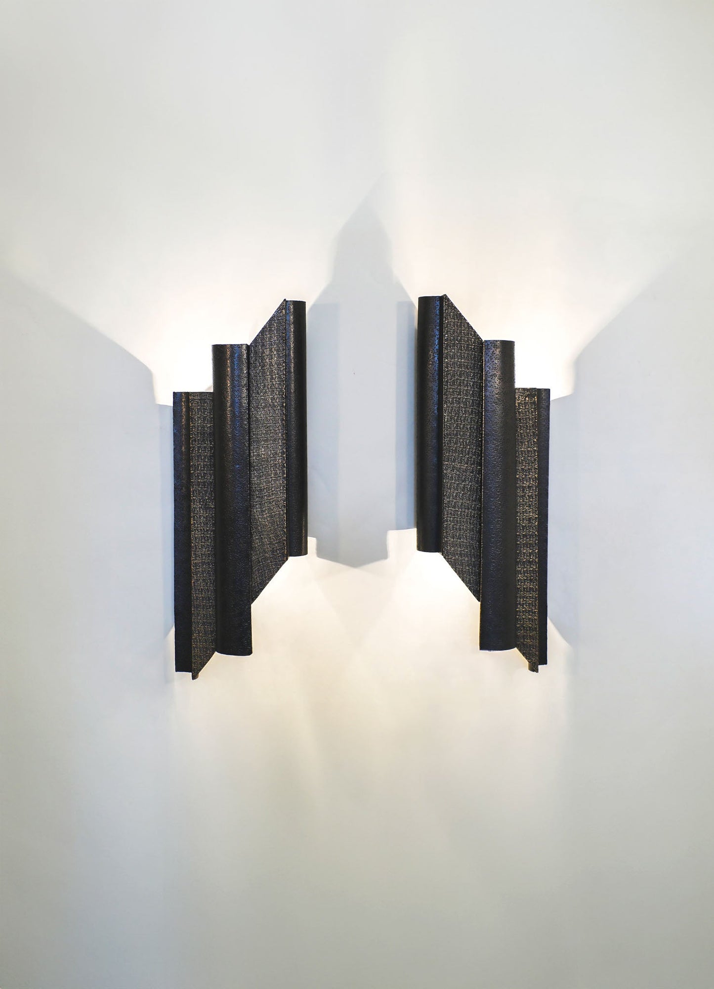 A modern wall sconce featuring a black textured metal design with geometric, layered panels, casting light to create an intricate pattern of shadows on a white wall.