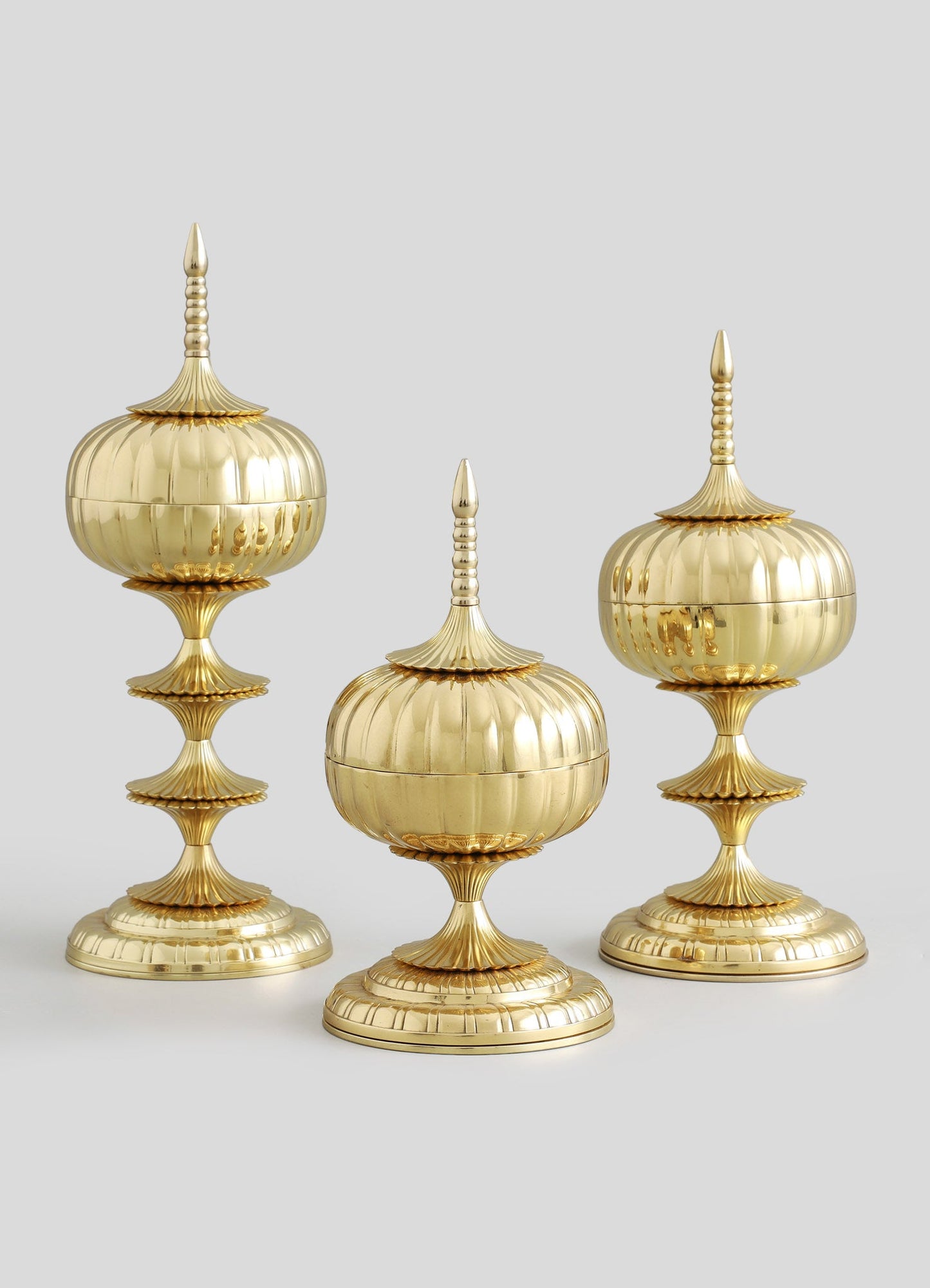 Three gold decorative finials with polished metal finish, featuring ribbed and scalloped motifs, mounted on stepped bases with ornate detailing.