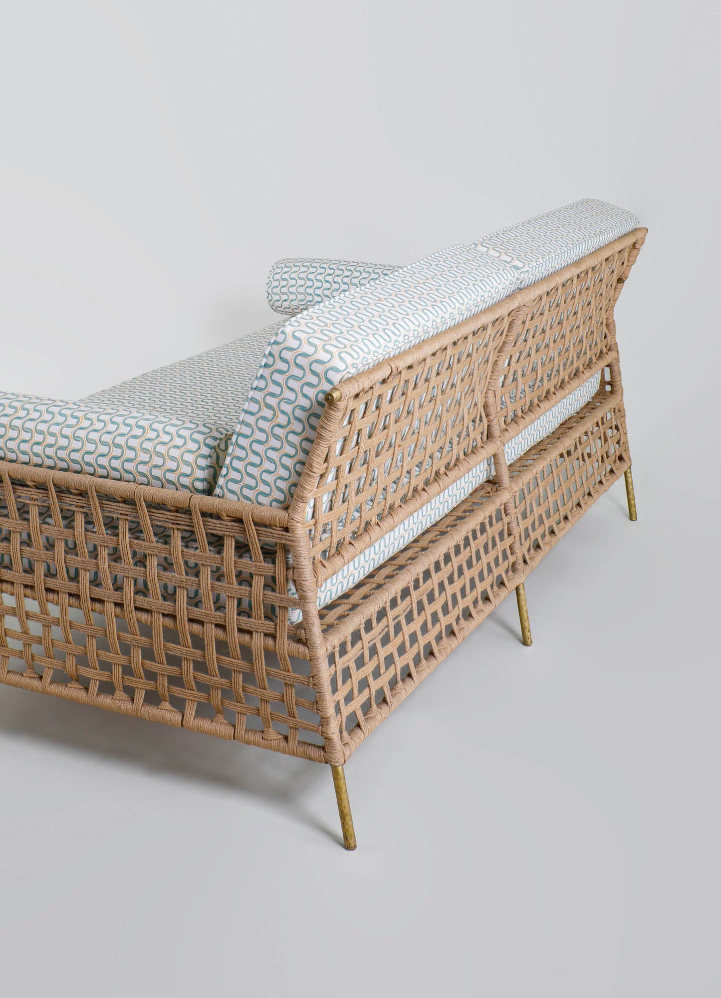 Wicker daybed with a natural rattan frame featuring woven patterns and tapered metal legs, dressed with a patterned fabric cushion and bolster in light tones.