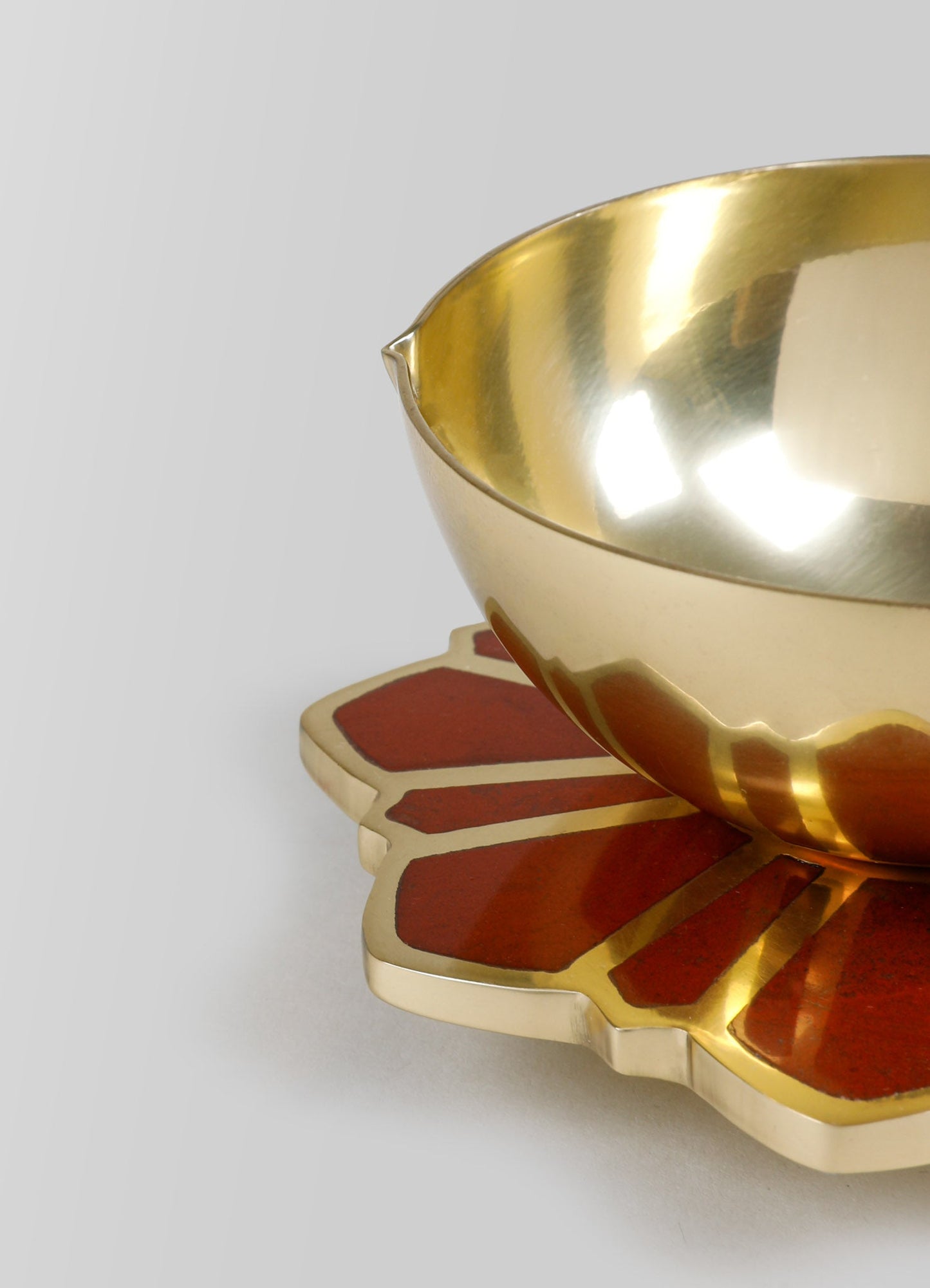 Brass bowl with a polished, reflective gold finish, placed on a decorative wooden and brass tray with a floral motif in red and beige tones. The design features smooth curves and a blend of metallic and painted elements, reflecting a contemporary and artisanal style.