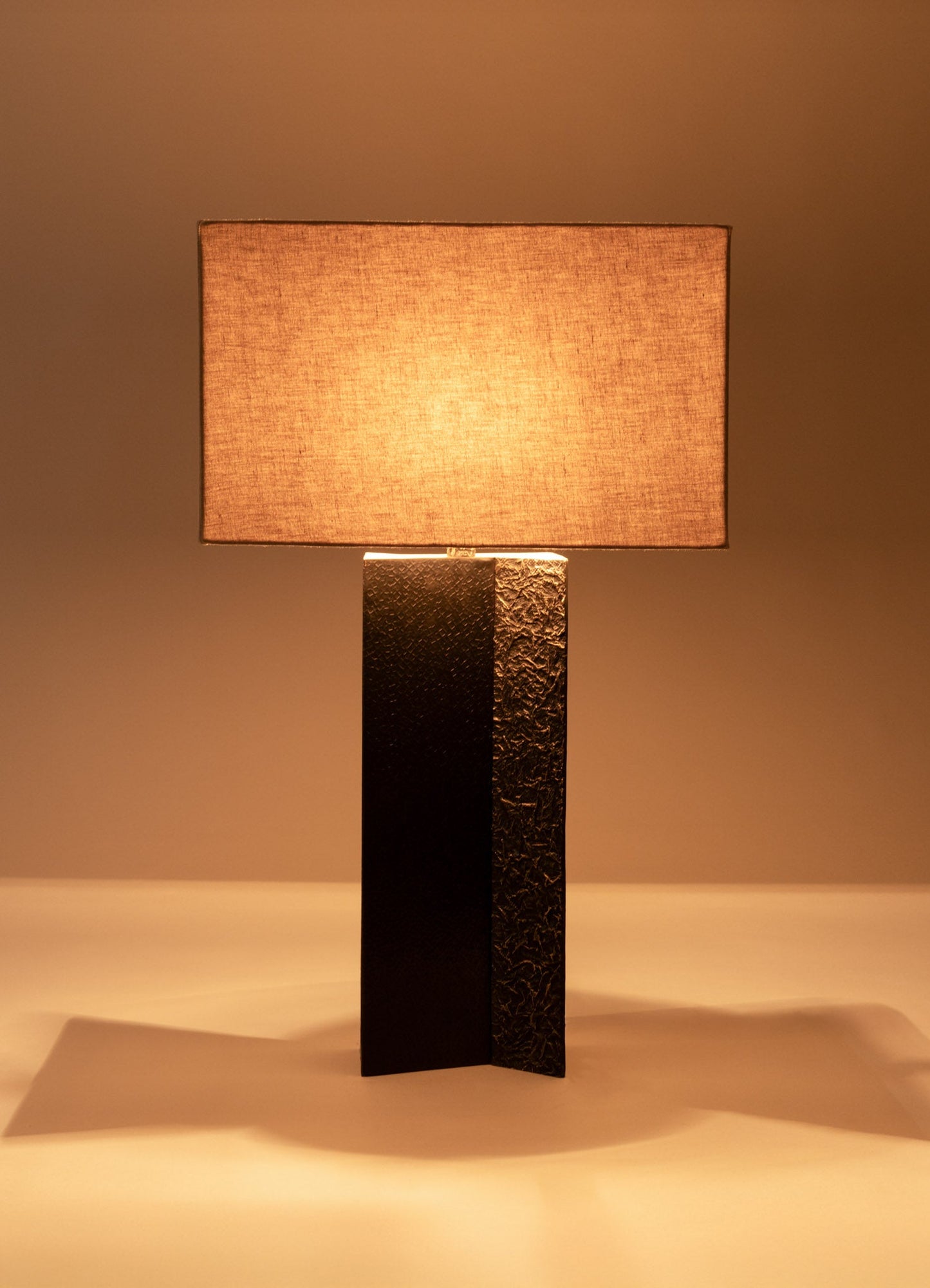 Table lamp with a rectangular fabric shade in a warm beige tone and a textured black metal base, emitting soft ambient light.