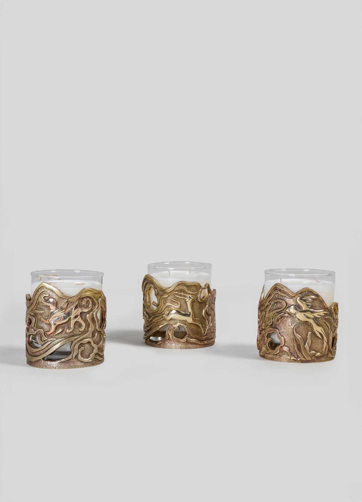 Three glass candles with ornate, openwork bronze metal holders featuring intricate, swirling motifs. The candles are arranged in a row against a plain, light-colored background.