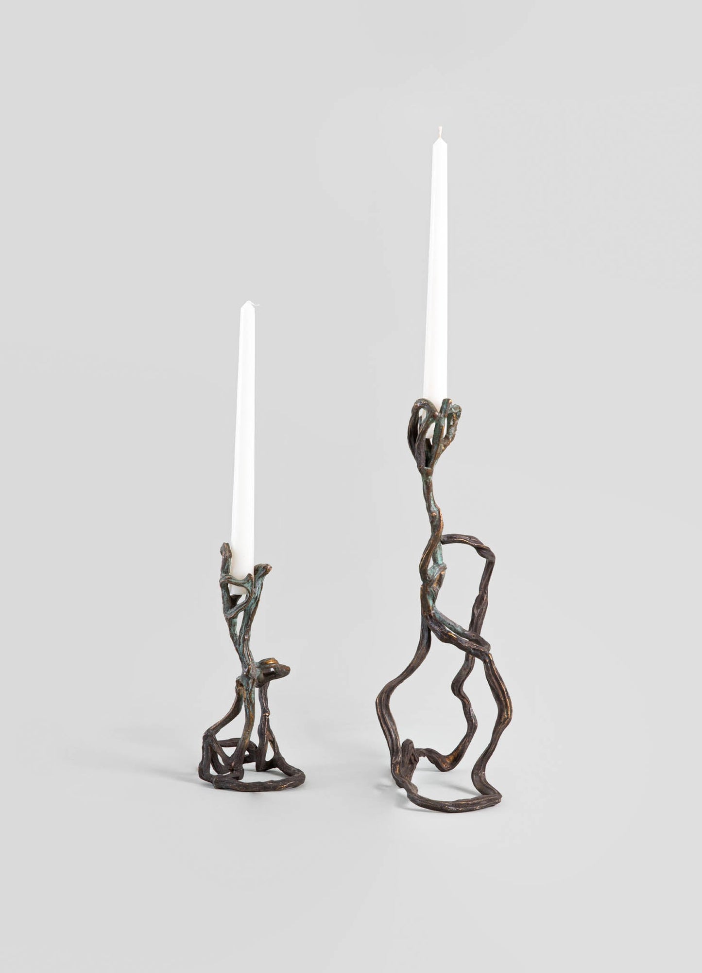 Brass candle holders with organic, twisted designs, featuring a dark, oxidized finish, holding tall white candles. The style is contemporary with handcrafted, sculptural motifs.