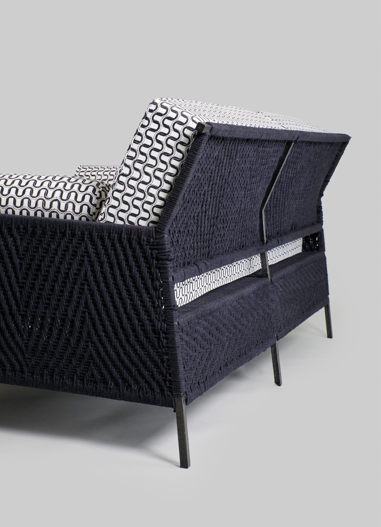 A modern armchair with a black woven rattan frame, featuring a sleek black metal base and legs, with black and white geometric patterned cushions for contrast.