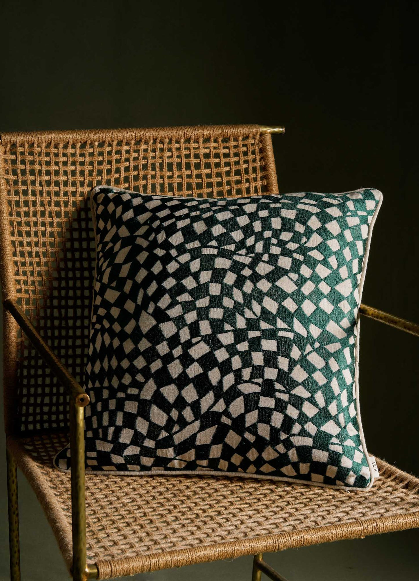 A decorative cushion with a black and white geometric pattern featuring concentric circles and squares, placed on a woven rattan chair with a natural finish. The background is dark, highlighting the contrast between the cushion's bold design and the chair's handcrafted texture.
