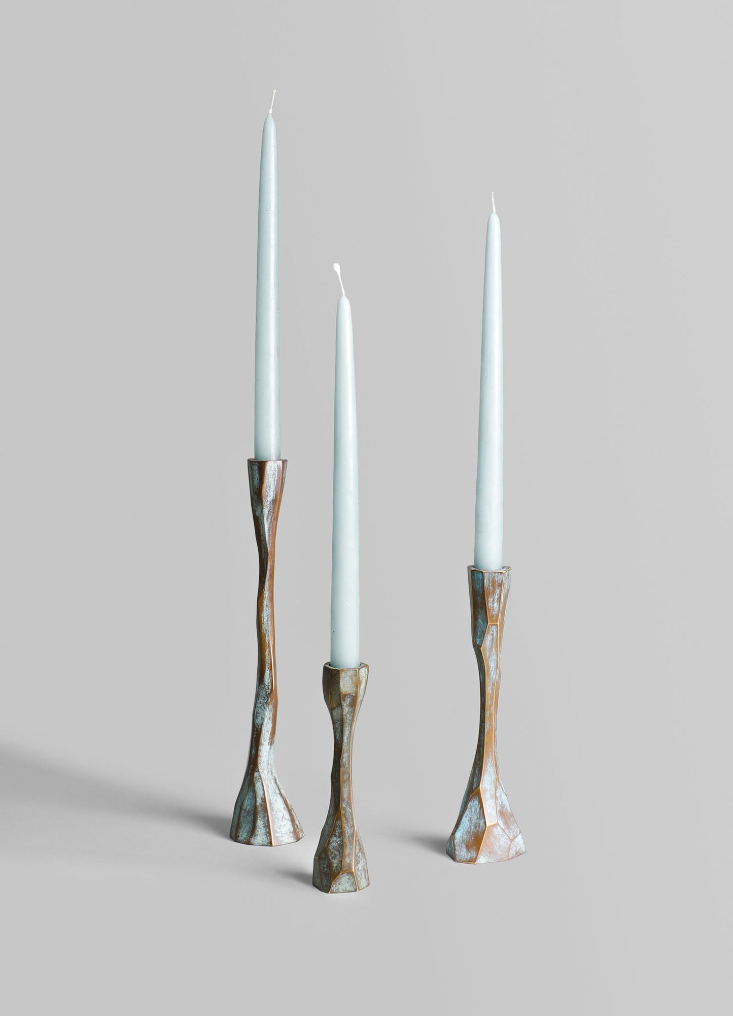 Three tall taper candles with white wax and unlit wicks are held in sculptural candle holders made of wood with a distressed, metallic finish in bronze and silver tones, set against a plain light gray background.