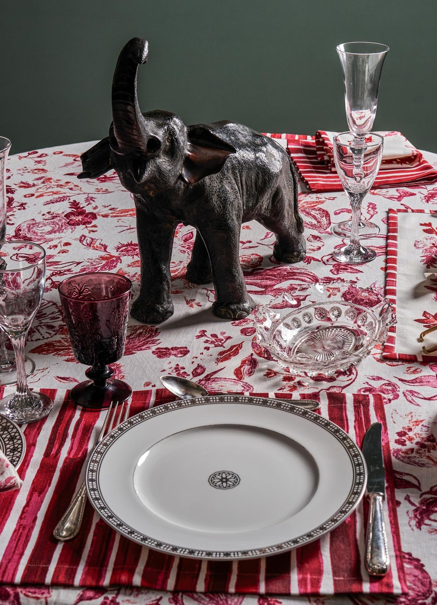 A black elephant figurine made of a glossy material serves as a decorative centerpiece on a dining table. The table is covered with a red and white floral-patterned tablecloth, complemented by clear and purple glassware, white and red napkins, and fine silverware.