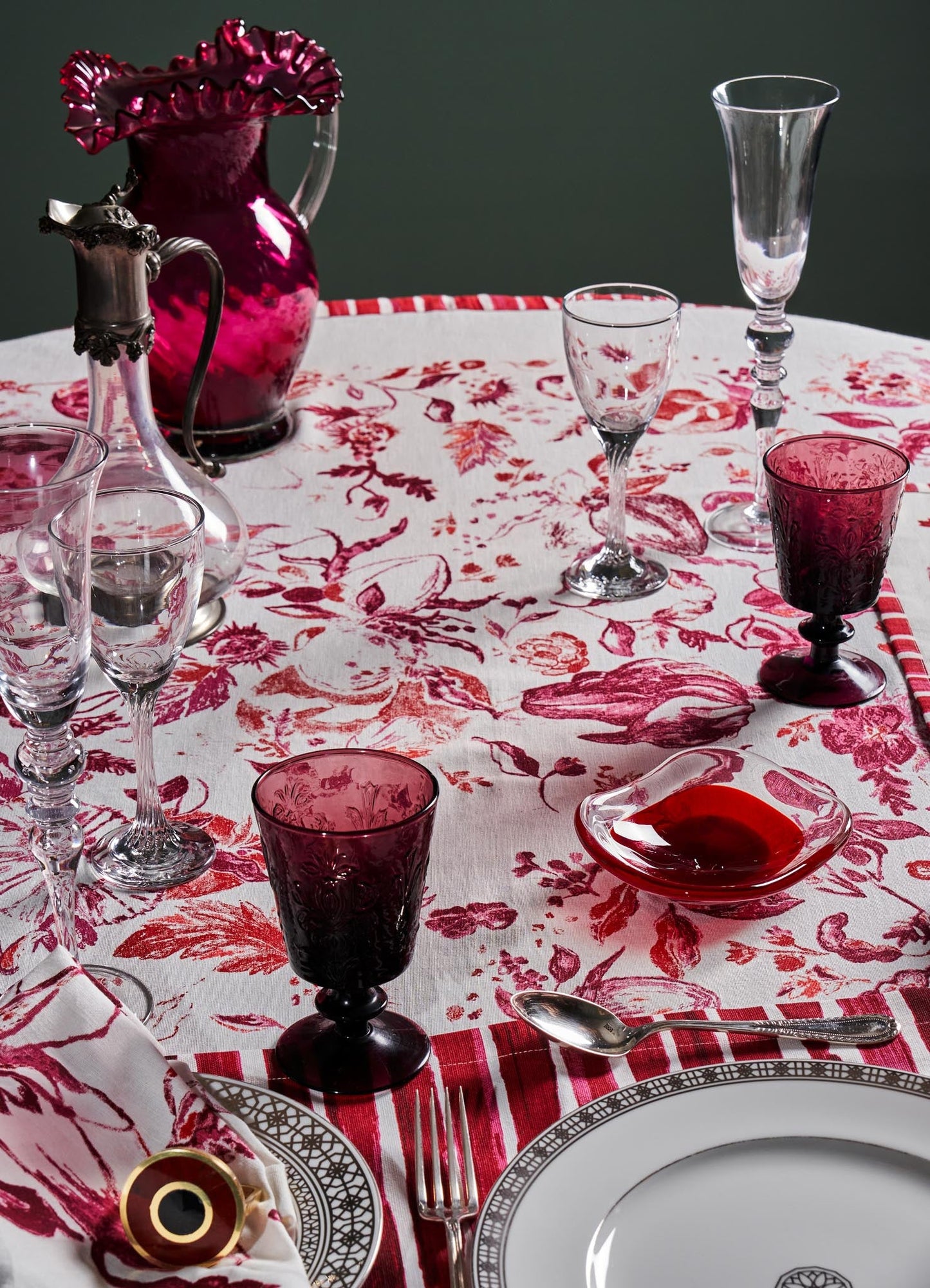 A dining table set with glassware including wine glasses and tumblers in shades of pink and clear, alongside a white tablecloth with red floral and botanical patterns, complemented by a porcelain plate with a black and gold geometric border, and silver cutlery.