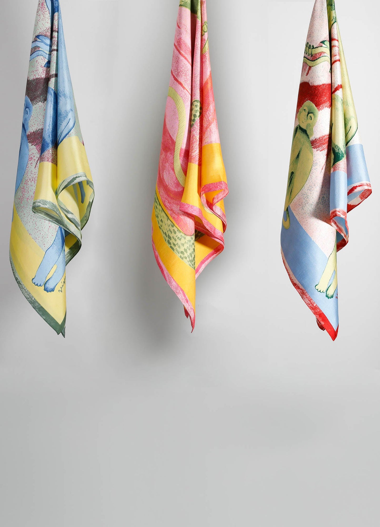 Four colorful tea towels with playful illustrations of animals and abstract motifs, featuring shades of blue, pink, yellow, and green, made from cotton or linen fabric, hung against a white background.