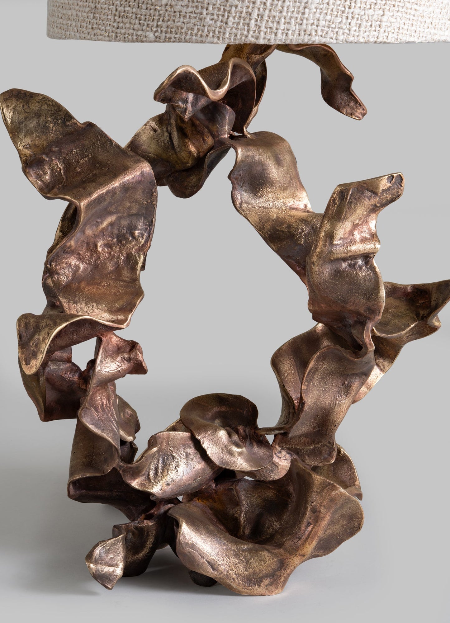 A decorative table lamp featuring a base made of abstract, organically shaped metallic copper with a textured finish, supporting a neutral fabric lampshade.