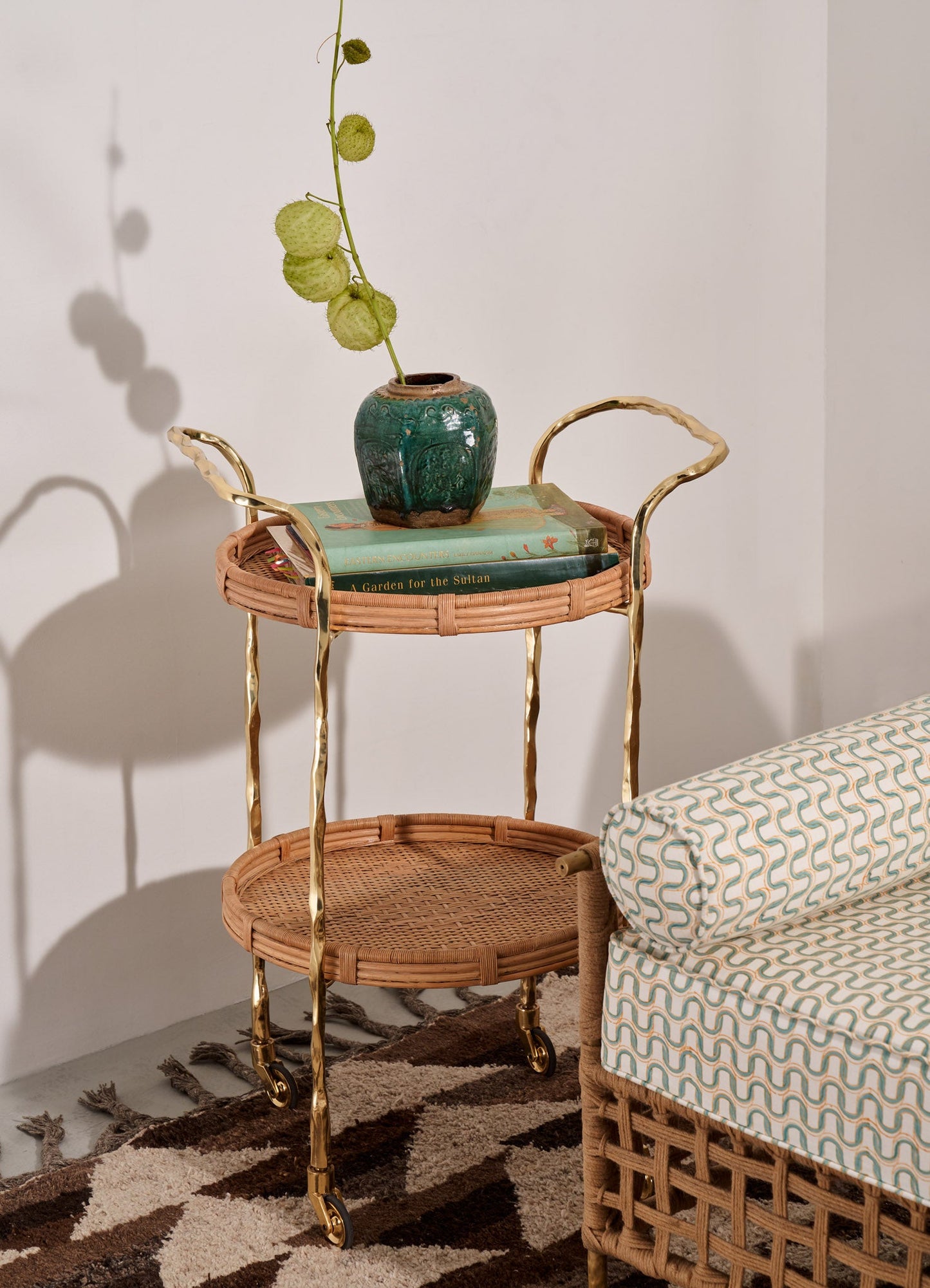 A three-tiered serving cart with a rattan frame and gold-toned metal handles and legs, featuring two round rattan shelves on casters. The scene includes a green ceramic vase with textured surface and spherical botanical accents, and a stack of books, set against a plain wall with a patterned rug on the floor.