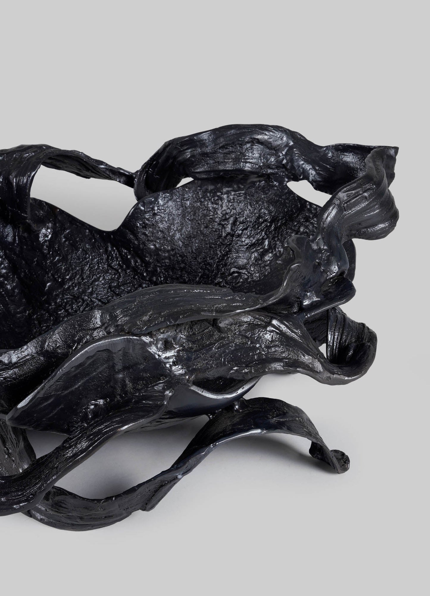 Abstract sculptural artwork composed of twisted, textured black material with a glossy finish. The piece has organic, flowing contours and a dynamic form against a plain, light grey background.