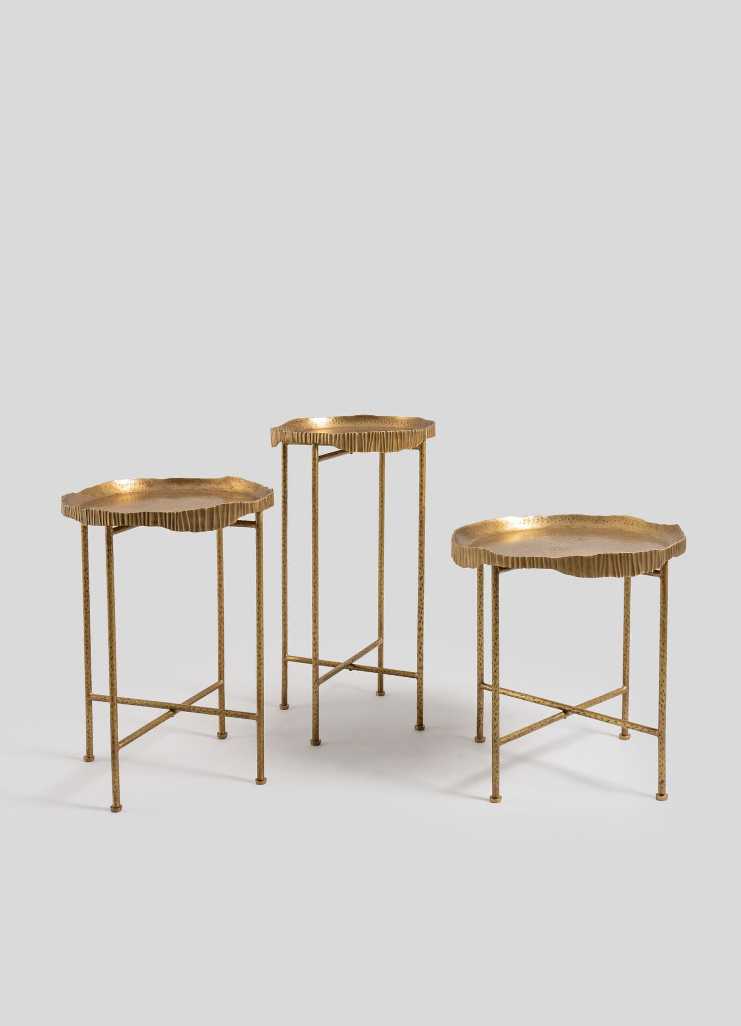 Three brass side tables with scalloped edges and textured surfaces, featuring slender legs with cross supports. The tables have a matte gold finish and a minimalist, contemporary style.