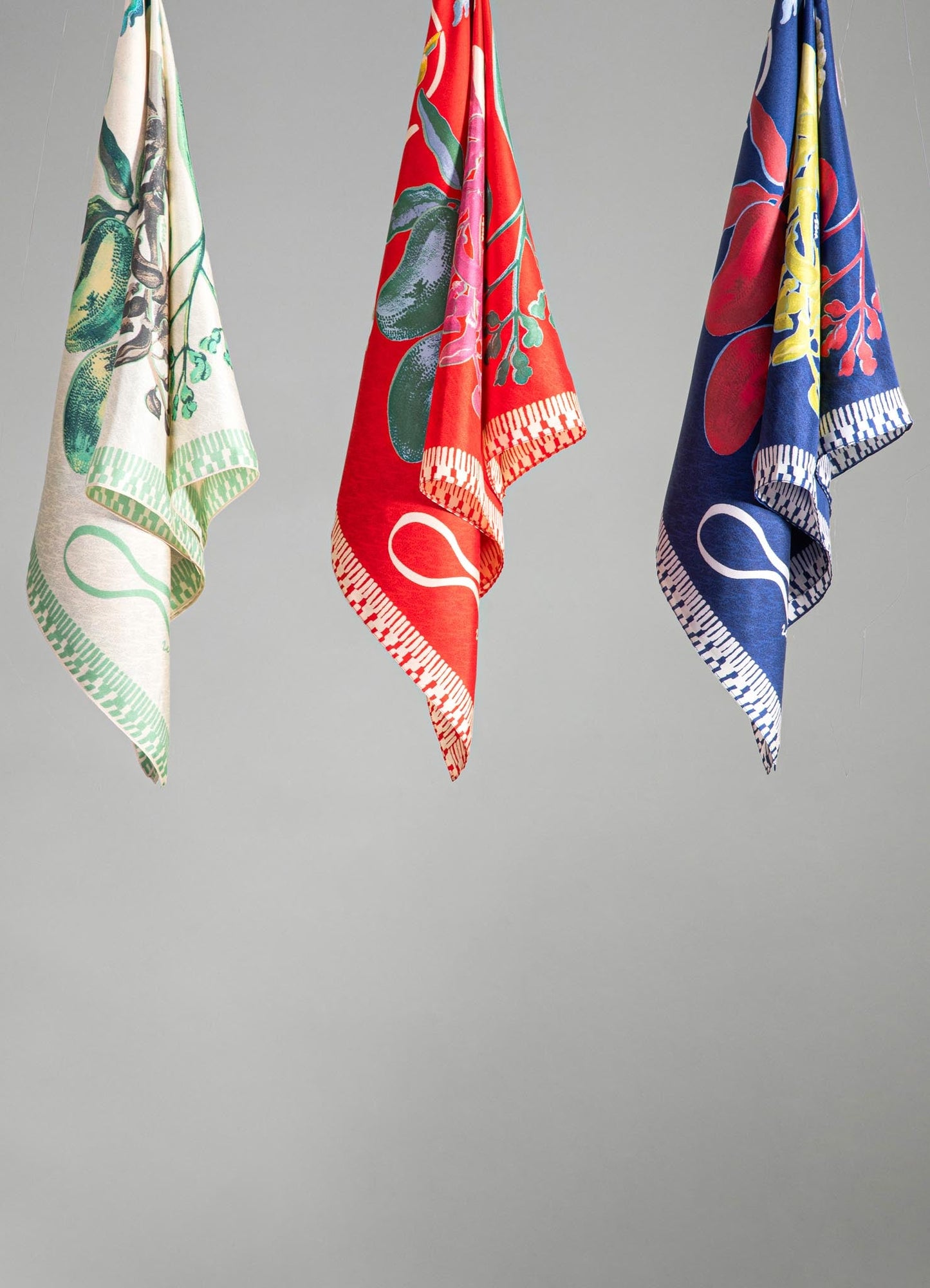 Softly draped fabric towels featuring vibrant floral patterns in red, green, and navy blue with white borders, hung against a gray background. The design style appears contemporary and lively with bold colors and botanical motifs.