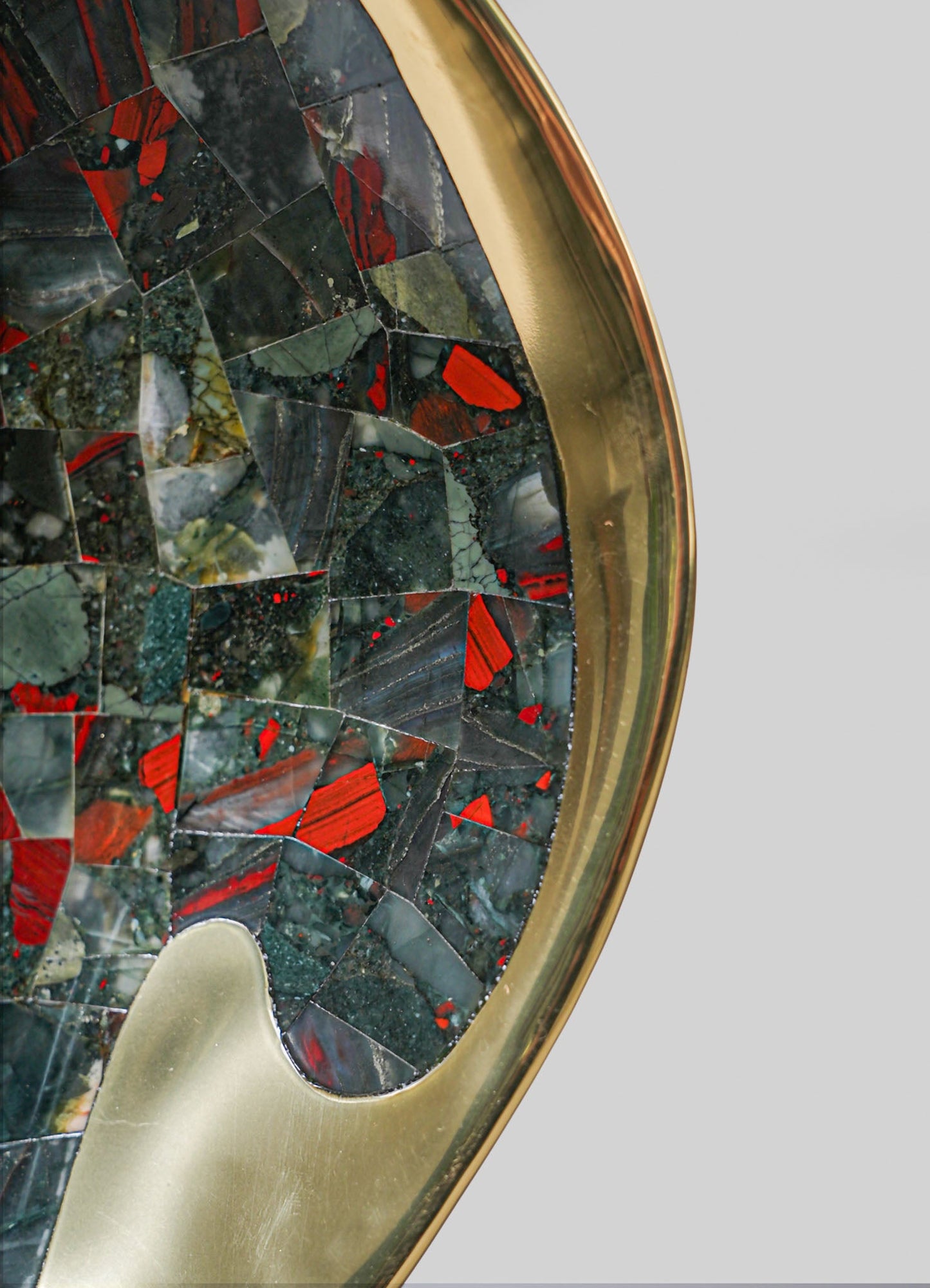 A round table with a metallic gold finish and a glass top featuring a mosaic pattern of multicolored stones in shades of black, red, green, and gray. The table has a contemporary style with an elegant, polished surface.