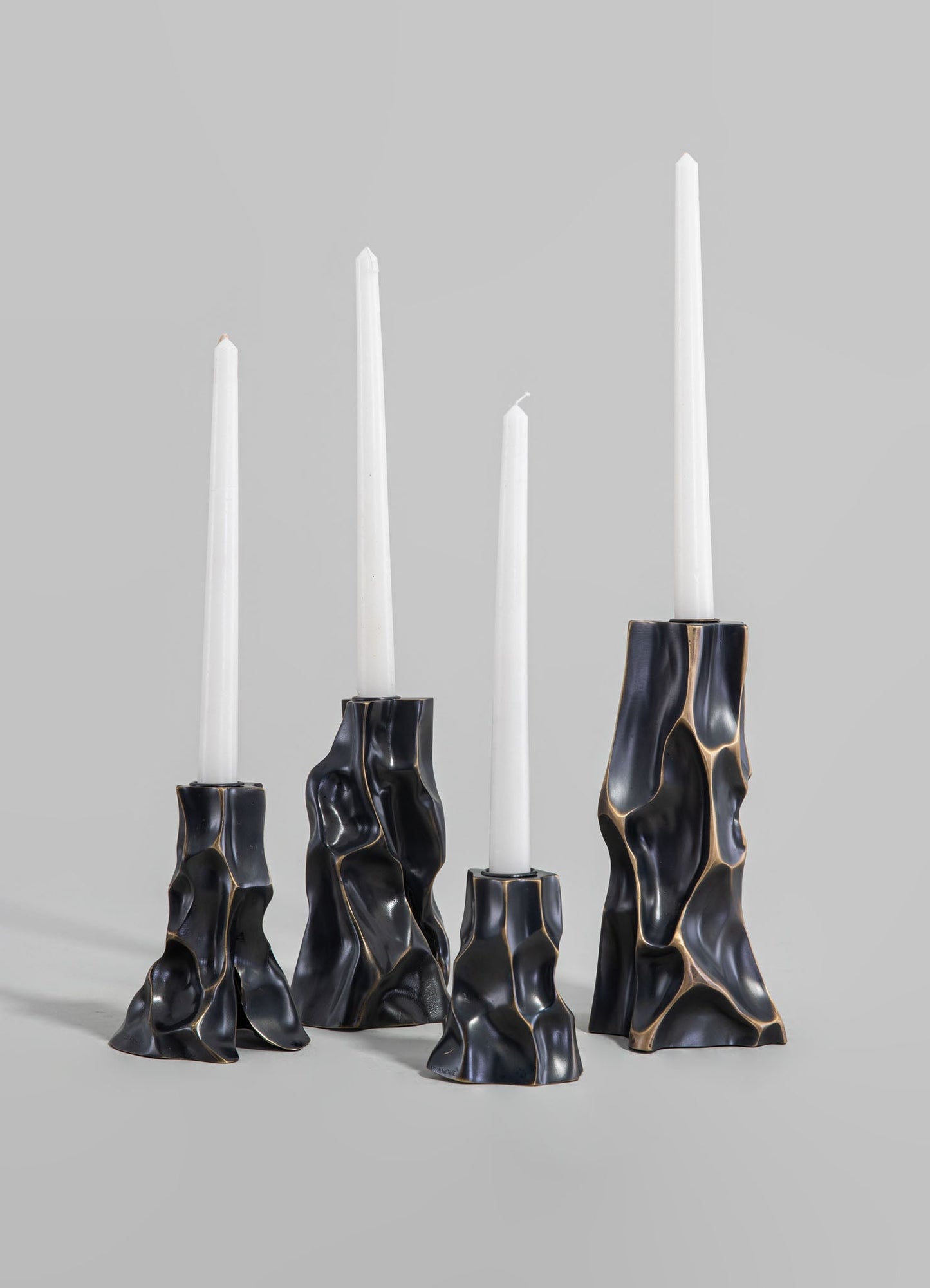 Set of four black sculptural candle holders with irregular, organic shapes and contrasting beige accents, designed to hold tall white tapered candles.