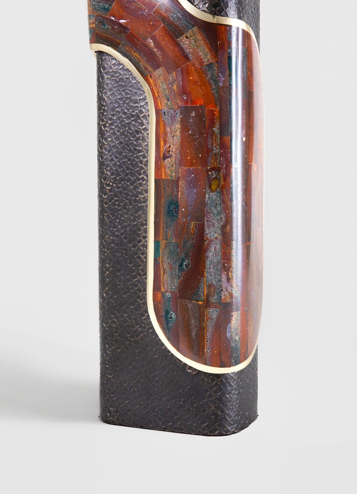 A tall decorative table or sculpture featuring a cylindrical shape, with a textured black surface and an inset panel of iridescent multicolored stone or glass with earthy tones, accentuated by a thin gold or brass trim.