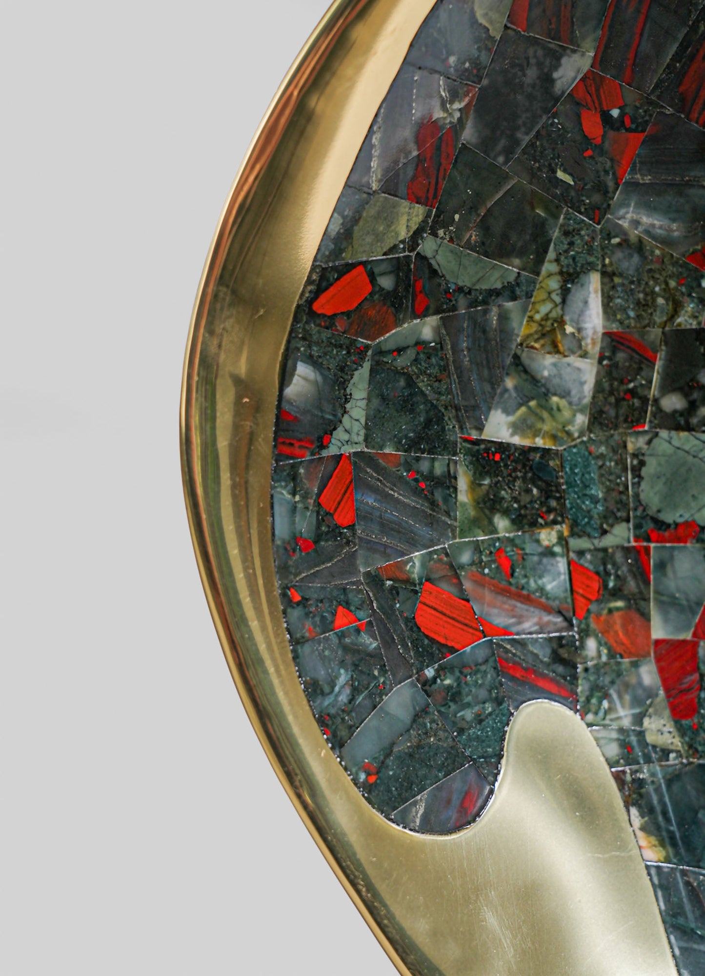 A close-up of a round tabletop featuring a mosaic of stone and glass with red, black, and gray tones, framed by a metallic gold edge. The style appears contemporary with a focus on bold, colorful motifs and a polished finish.