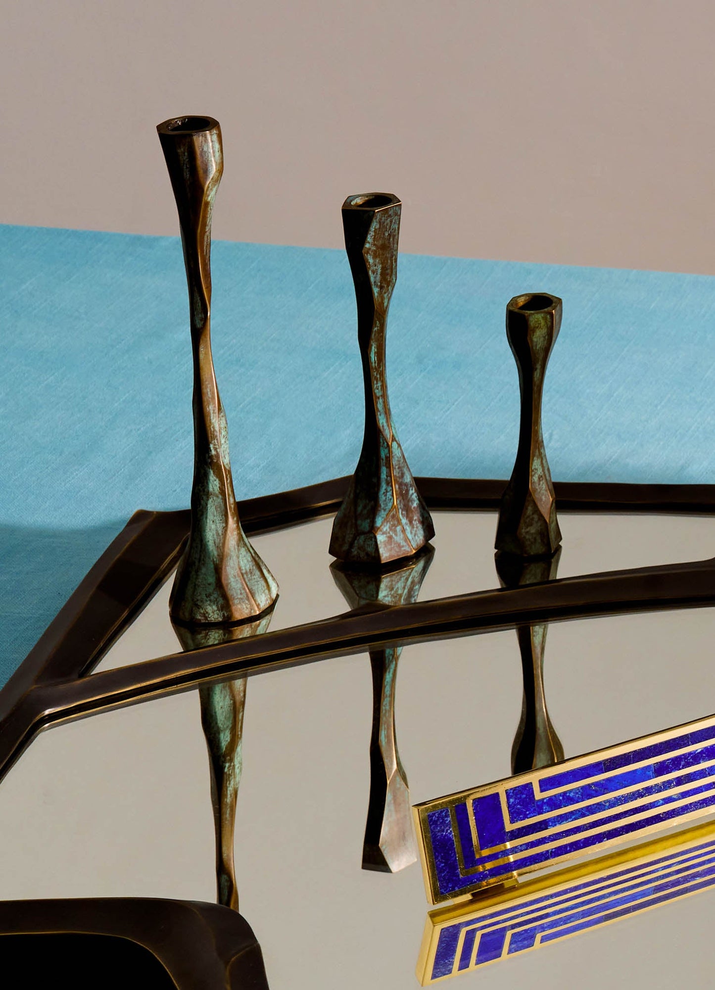 Brass candle holders with elongated, twisted forms in varying heights, featuring a patina finish. The set is placed on a reflective surface with a blue cloth background, emphasizing a contemporary style.